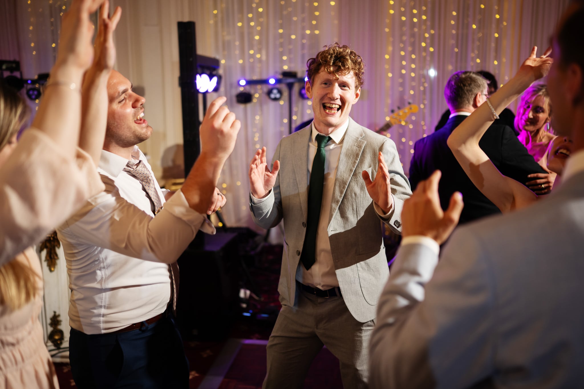 A group of people dressed in formal attire dance and smile at an indoor London wedding with string lights in the background at No. 4 Hamilton Place.