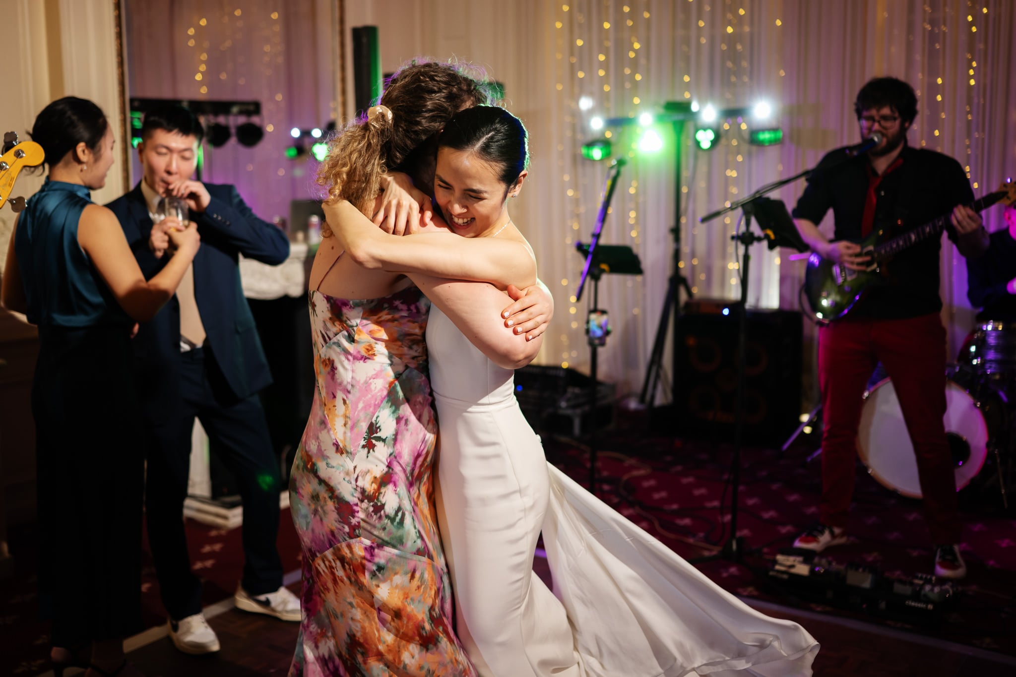 Two women embrace and dance joyfully at a London wedding celebration at No. 4 Hamilton Place, with a live band performing in the background as other guests enjoy the festive event.