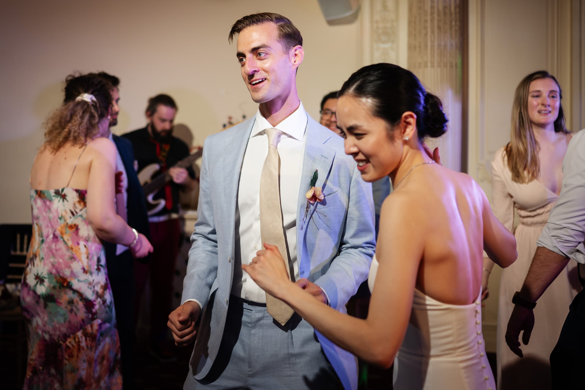 A man in a light blue suit and a woman in a white dress are dancing together at an indoor London wedding at No. 4 Hamilton Place, with other guests visible in the background.
