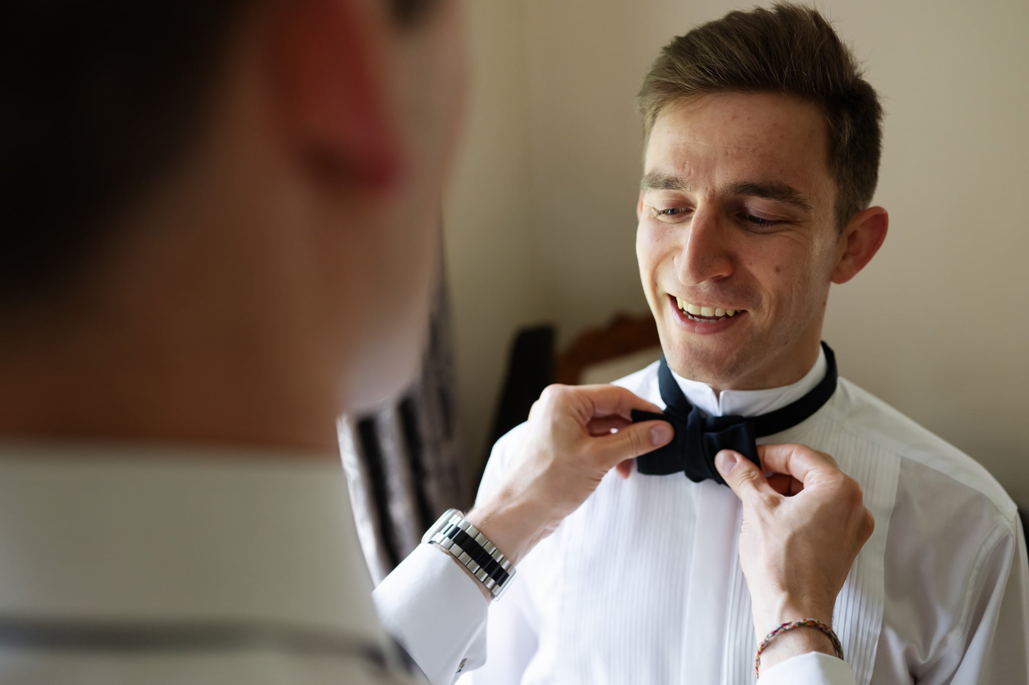 A man in a white shirt smiles while another person adjusts his black bow tie, capturing a joyful moment before the wedding.