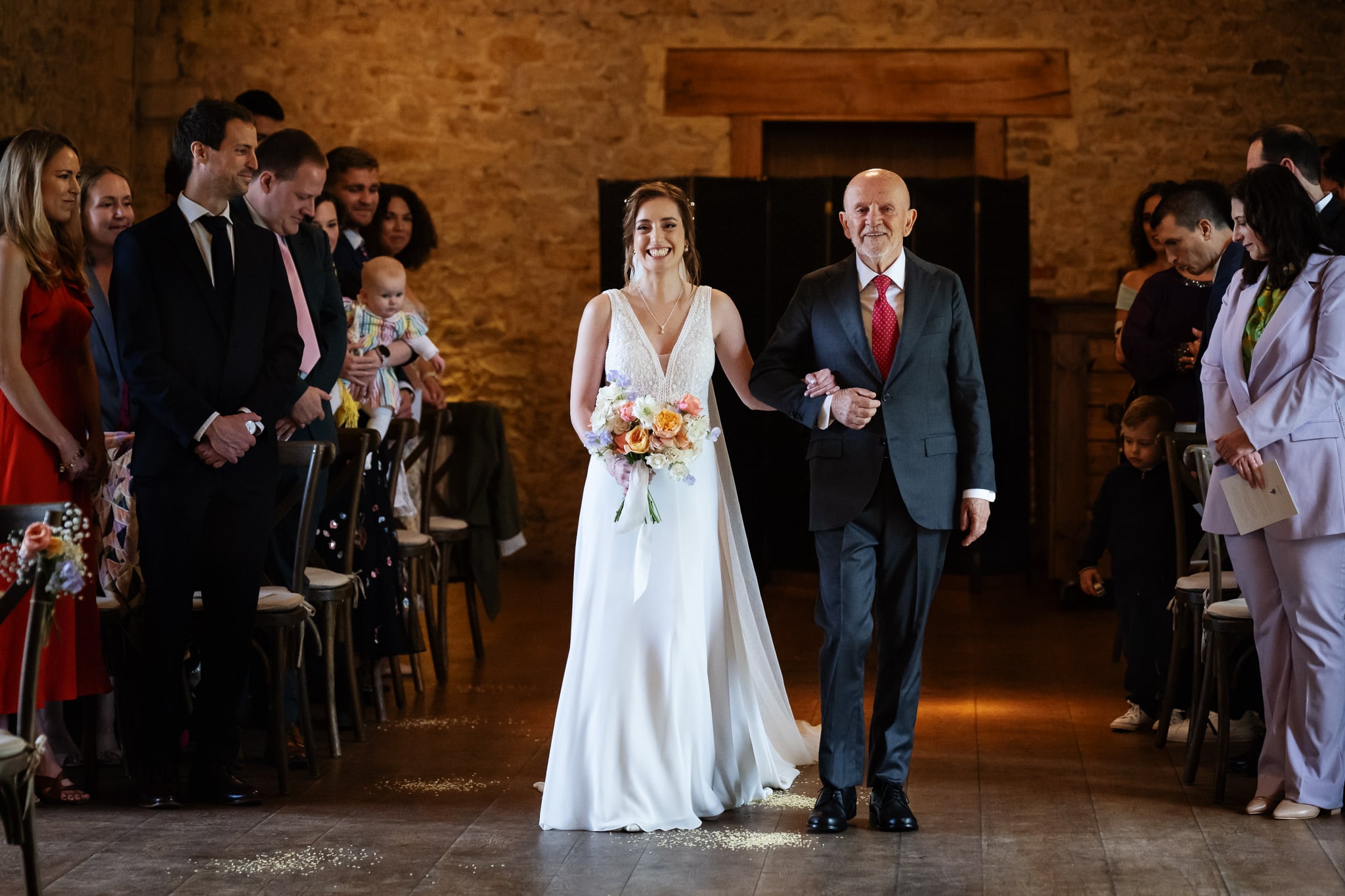 A bride in a white dress holding a bouquet walks down the aisle with an older man in a suit, surrounded by guests at her wedding in the rustic indoor venue of Notley Abbey.