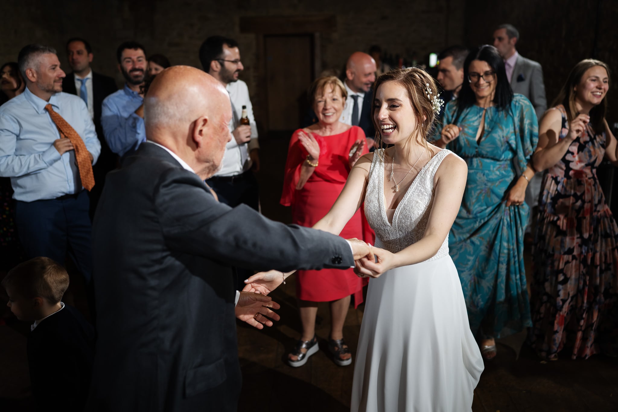 A bride in a white dress dances with an older man at a wedding reception, while the groom and smiling guests look on.