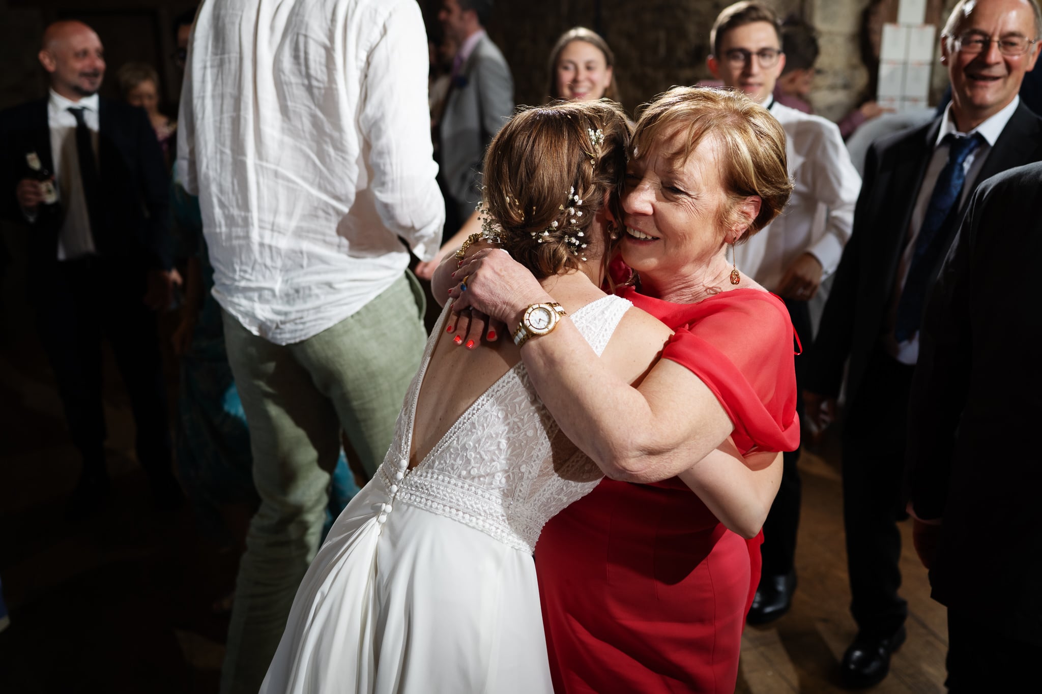 A bride in a white dress hugs a woman in a red dress, surrounded by smiling guests and the groom at a joyful wedding reception.