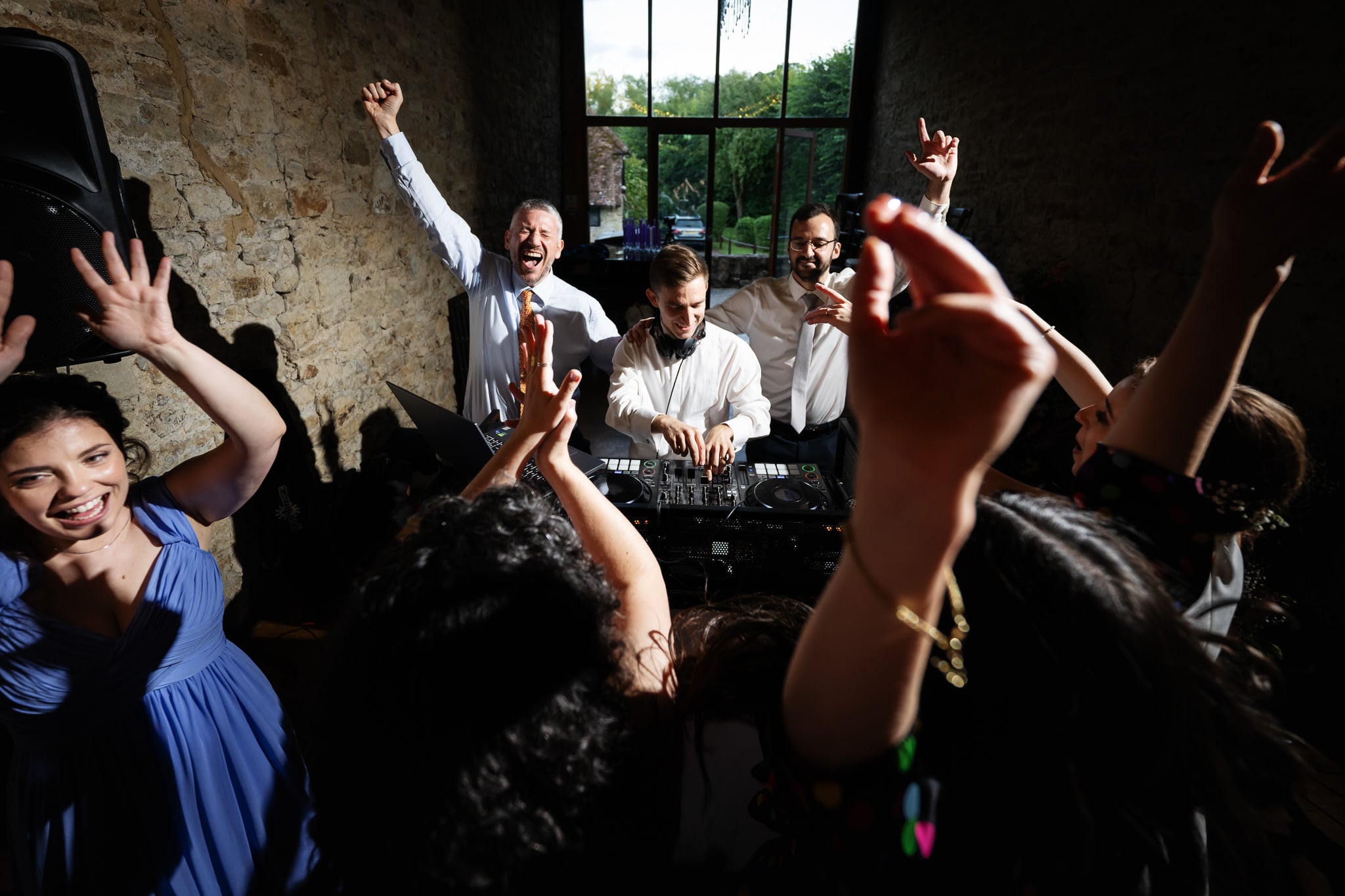 A group of people are dancing and raising their hands while a DJ plays music indoors near a large window at Notley Abbey in Buckinghamshire.