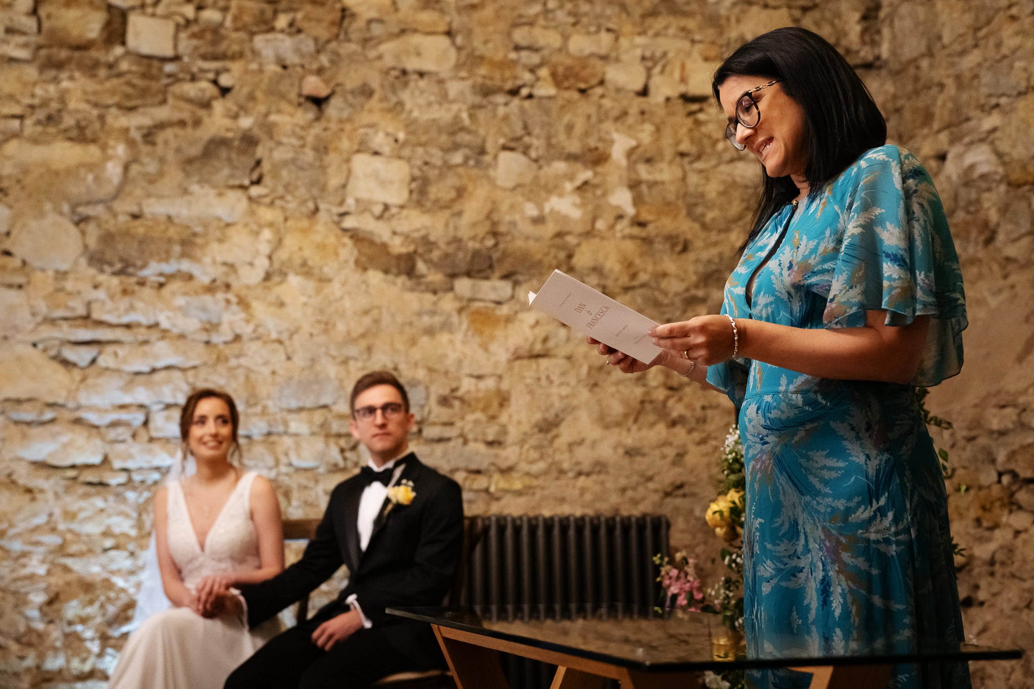 A woman in a blue dress reads from a card at a Buckinghamshire wedding, while the bride and groom sit together holding hands in front of a stone wall.