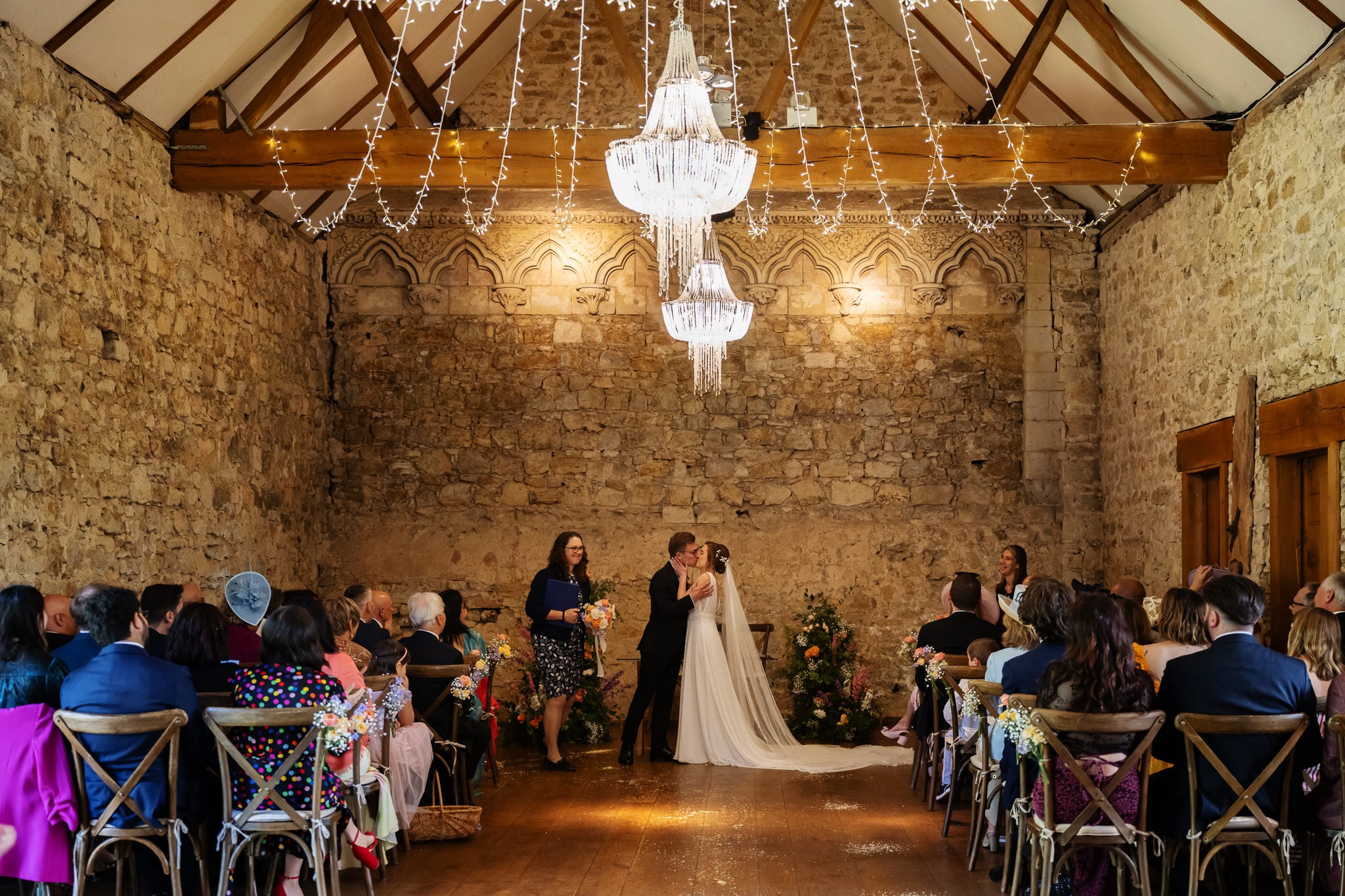 A couple shares their first kiss as newlyweds in a rustic stone hall in Buckinghamshire, surrounded by seated guests and glowing chandeliers.
