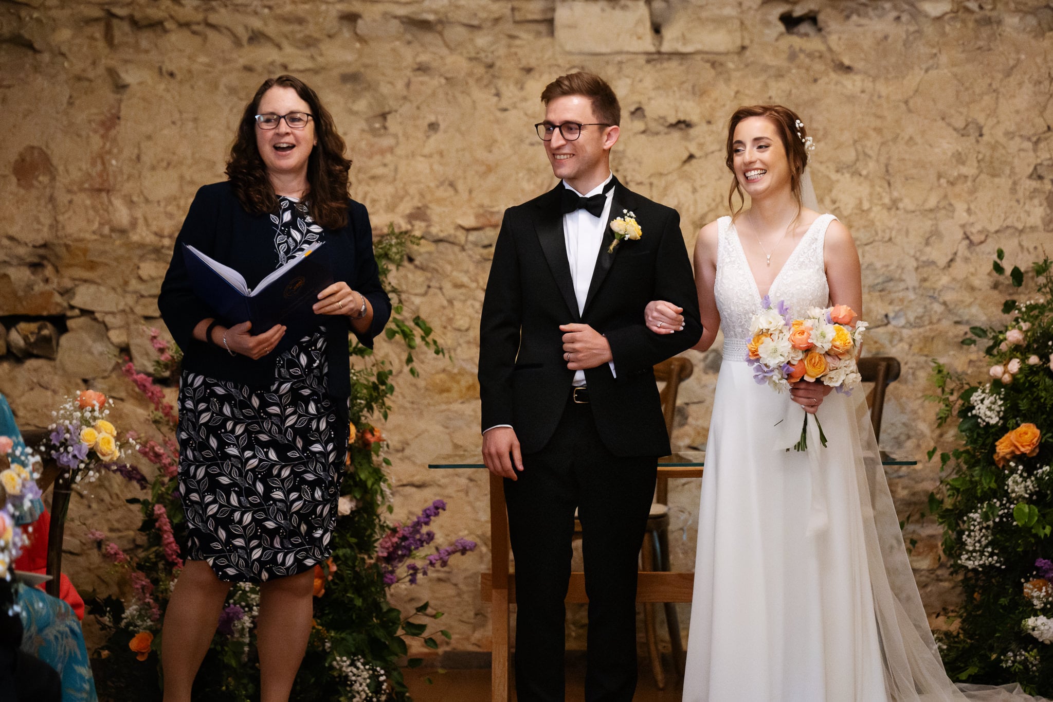 A newlywed couple stands arm in arm, smiling beside an officiant holding a book at Notley Abbey in Buckinghamshire. Indoors, they are surrounded by flowers and a charming stone wall background.