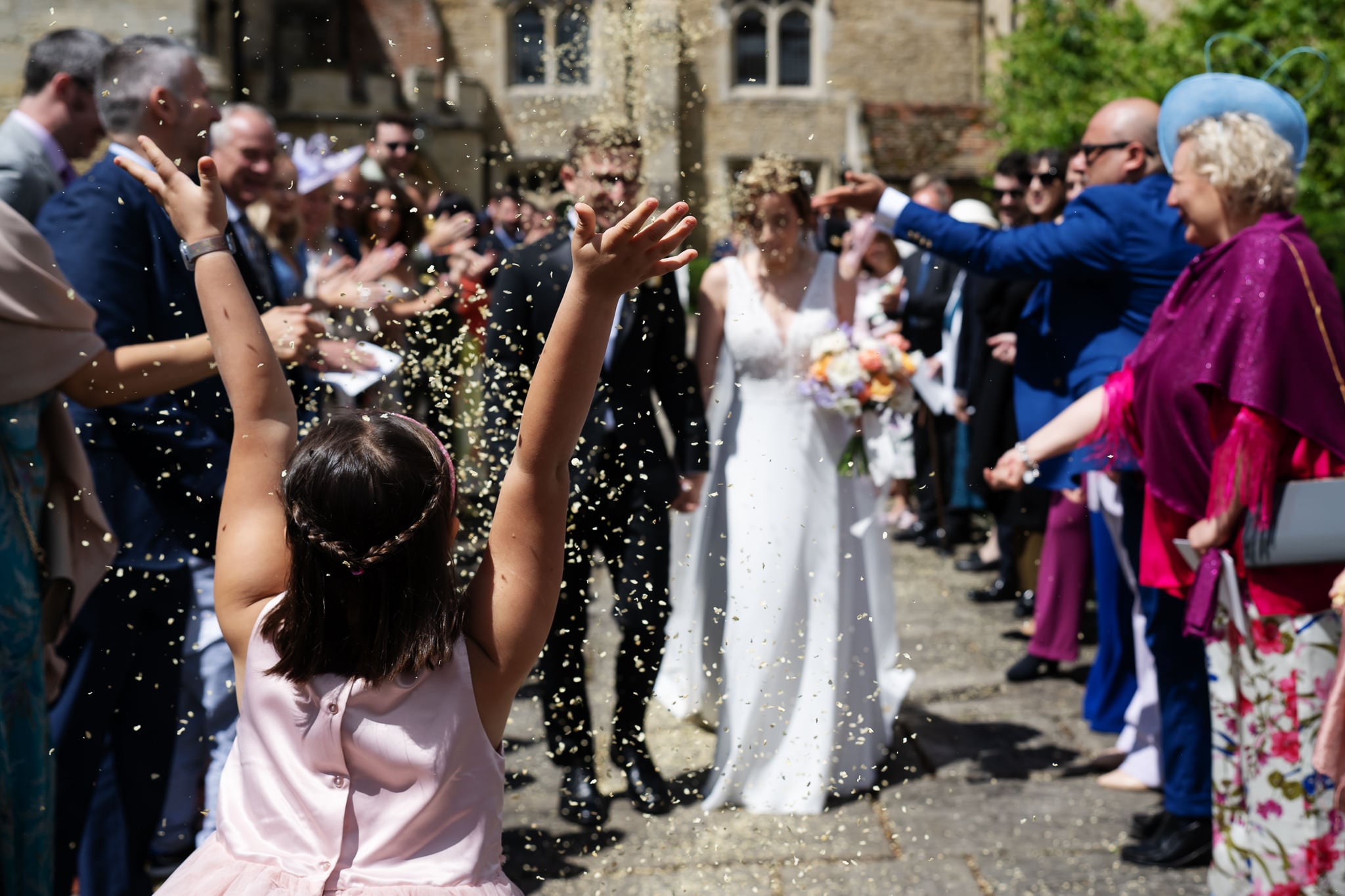 A girl throws confetti as a bride and groom walk down an outdoor aisle at Notley Abbey in Buckinghamshire, surrounded by cheering guests in formal attire.
