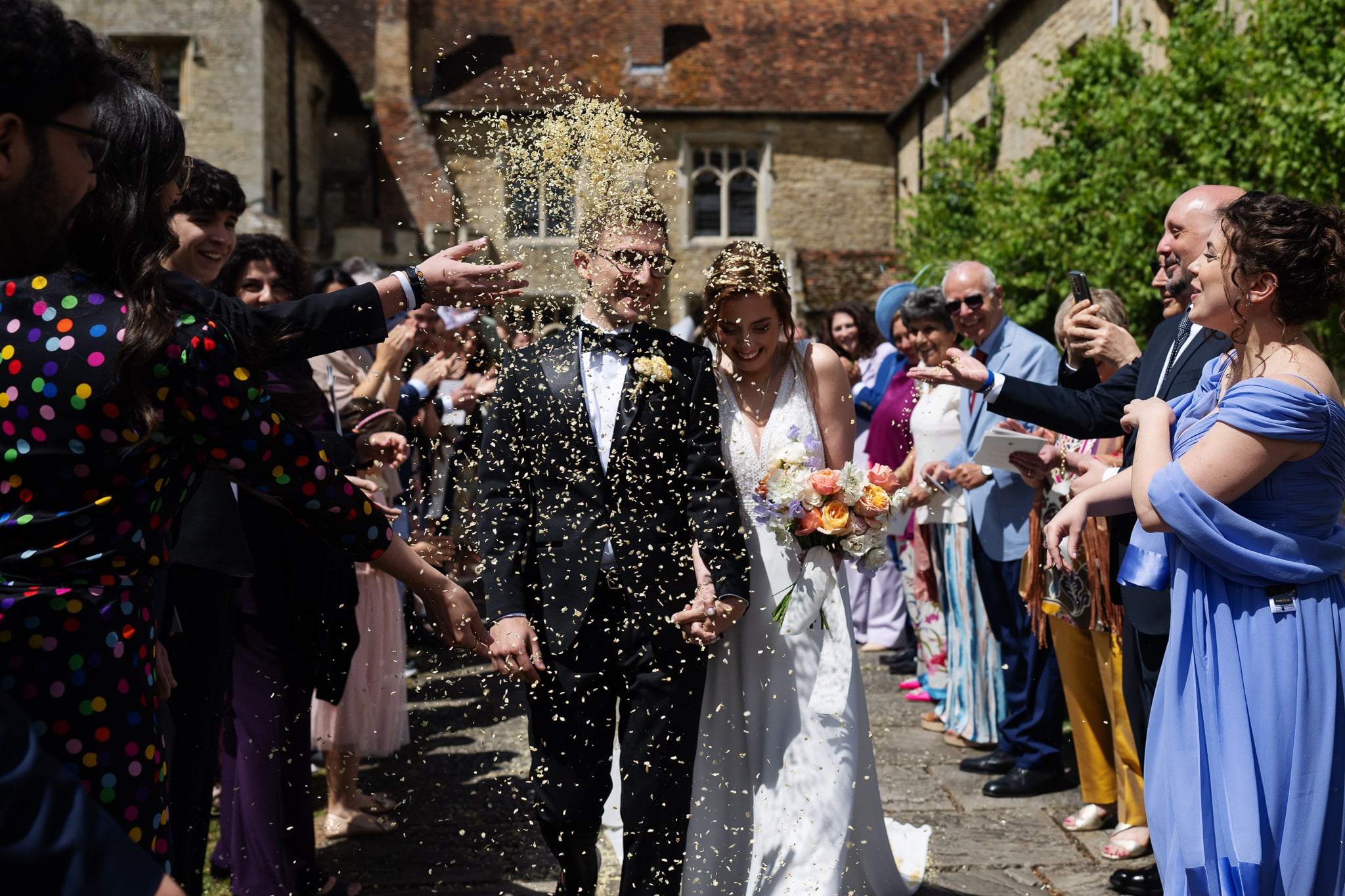 A newlywed couple walks outside their Buckinghamshire wedding as guests throw confetti; the smiling bride holds a bouquet amid a joyful, celebratory crowd.