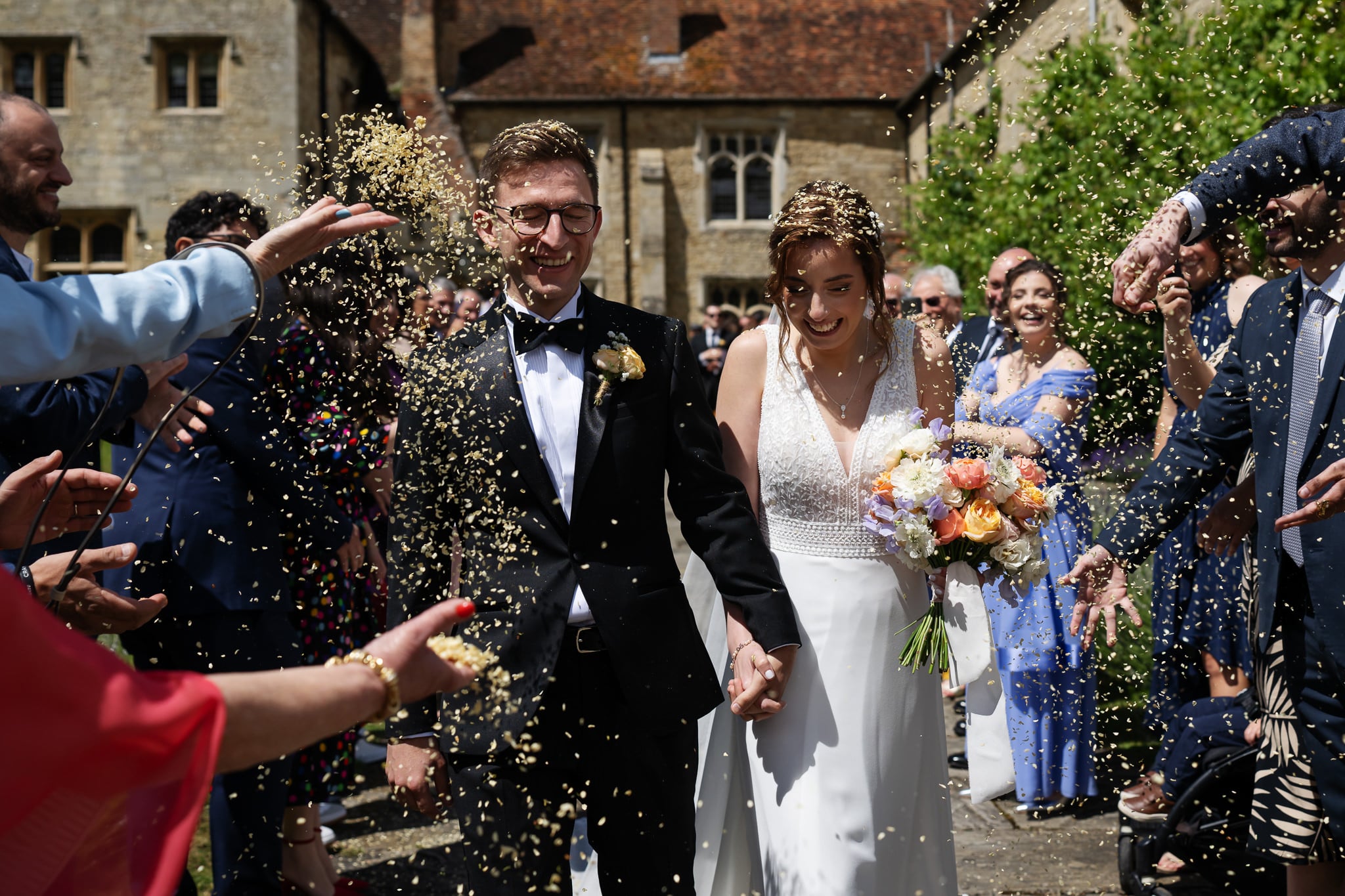 A bride and groom walk hand in hand outdoors at Notley Abbey while guests throw confetti at them. The bride holds a colorful bouquet as people smile and celebrate around them.