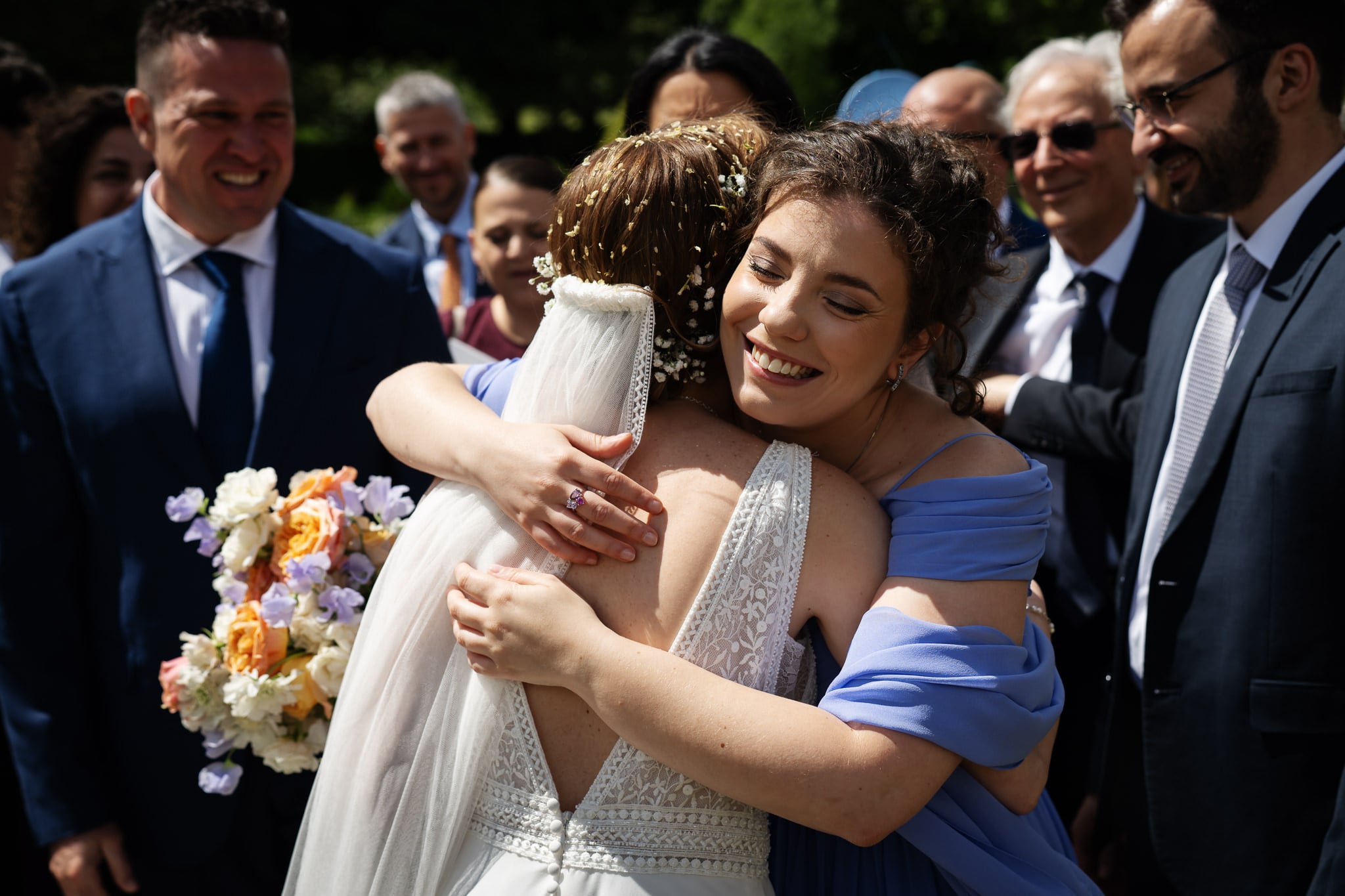 A bride and a woman in a blue dress hug outdoors at a Buckinghamshire wedding, surrounded by smiling guests. The bride holds a bouquet of flowers.