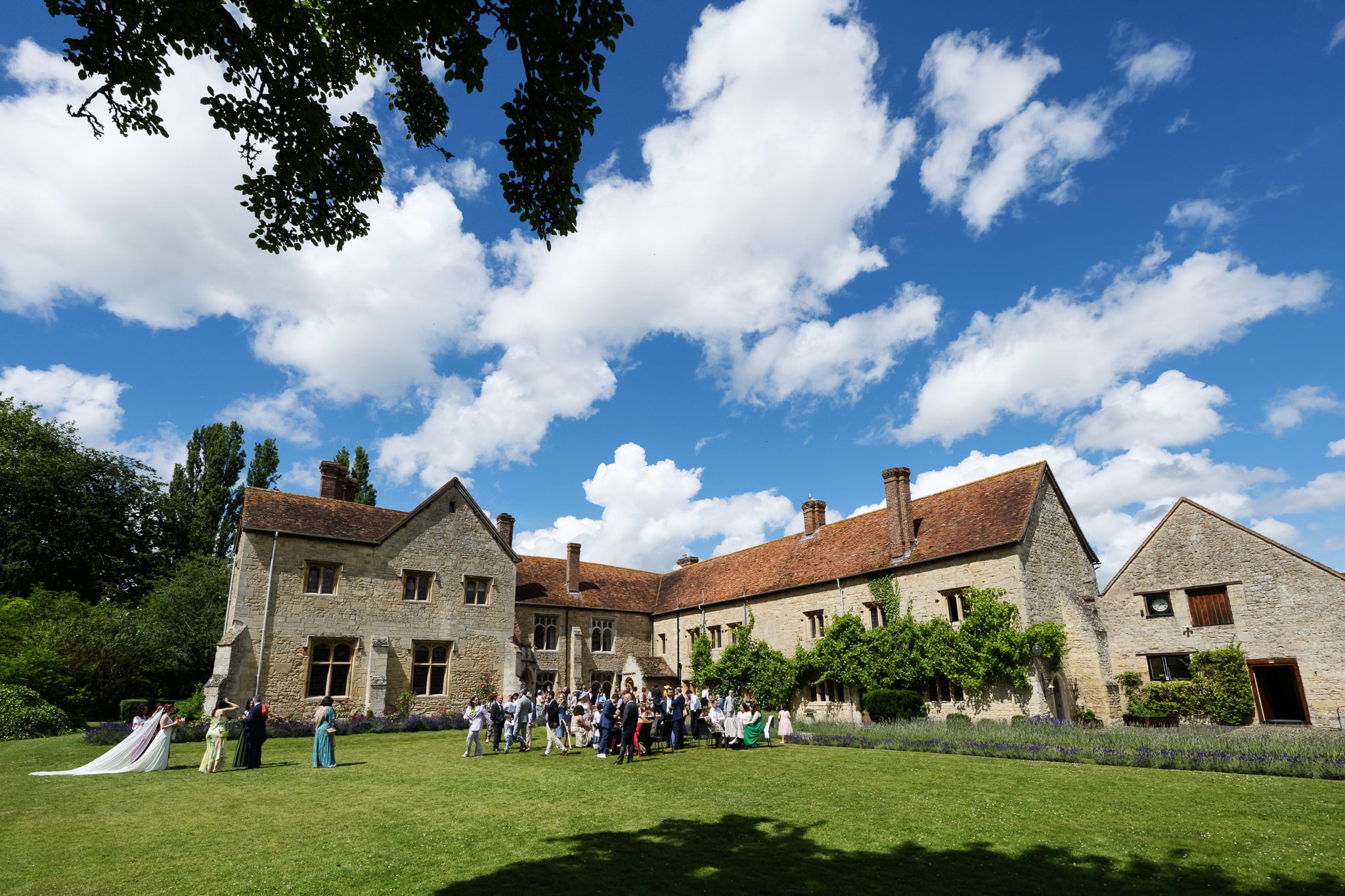 A group of people gathers on the lawn in front of Notley Abbey, a large, historic stone building with a red-tiled roof in Buckinghamshire, under a partly cloudy sky.