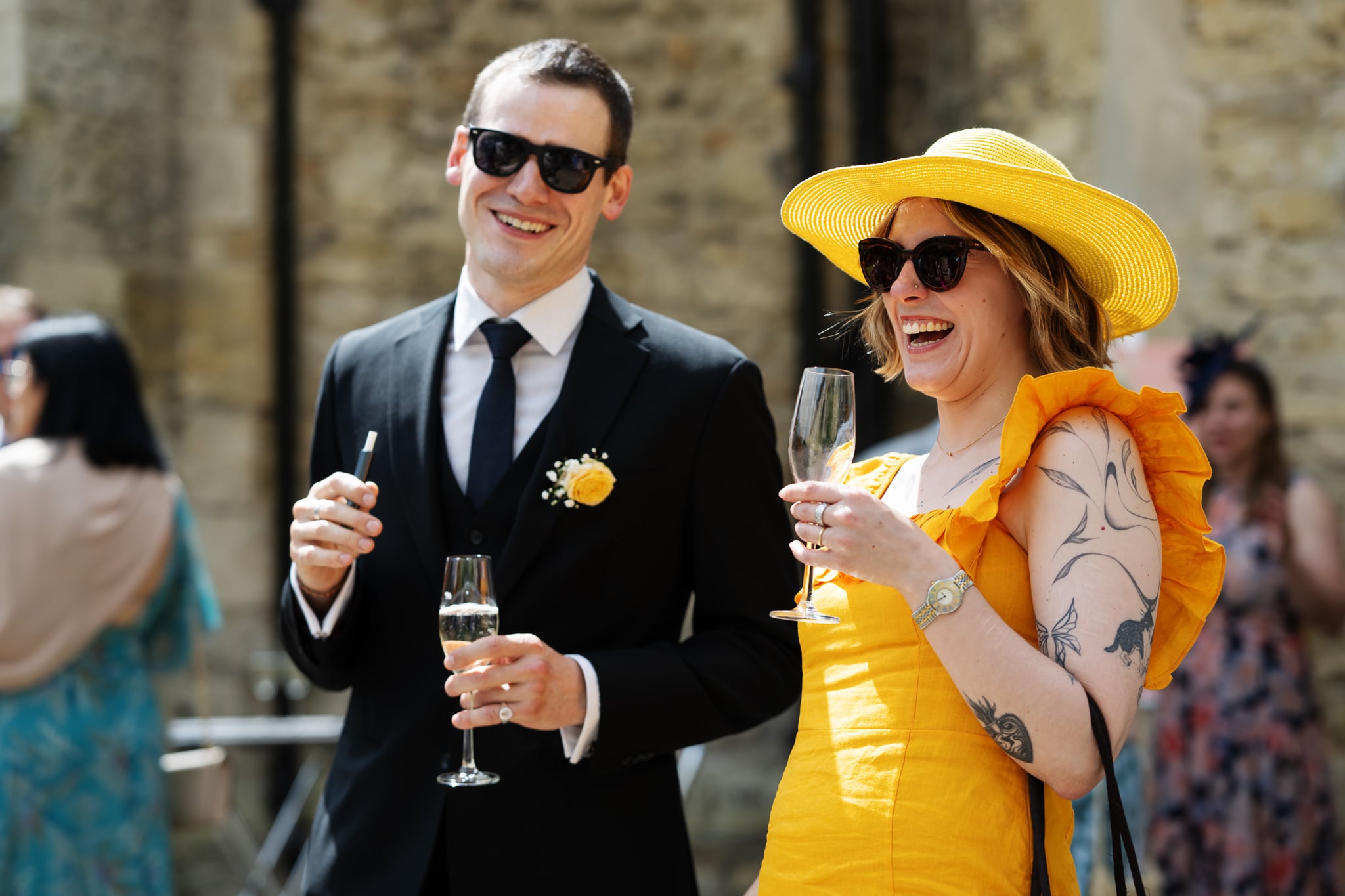 A man in a black suit and a woman in a yellow dress and hat, both wearing sunglasses, smile and hold drinks at an outdoor wedding celebration at Notley Abbey.