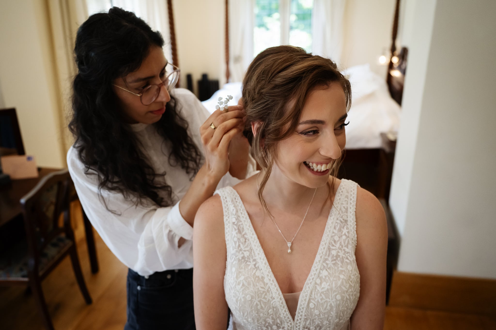 A bride in a white dress smiles while another woman adjusts her hair, as a photographer captures the moment in a well-lit room with a bed in the background.