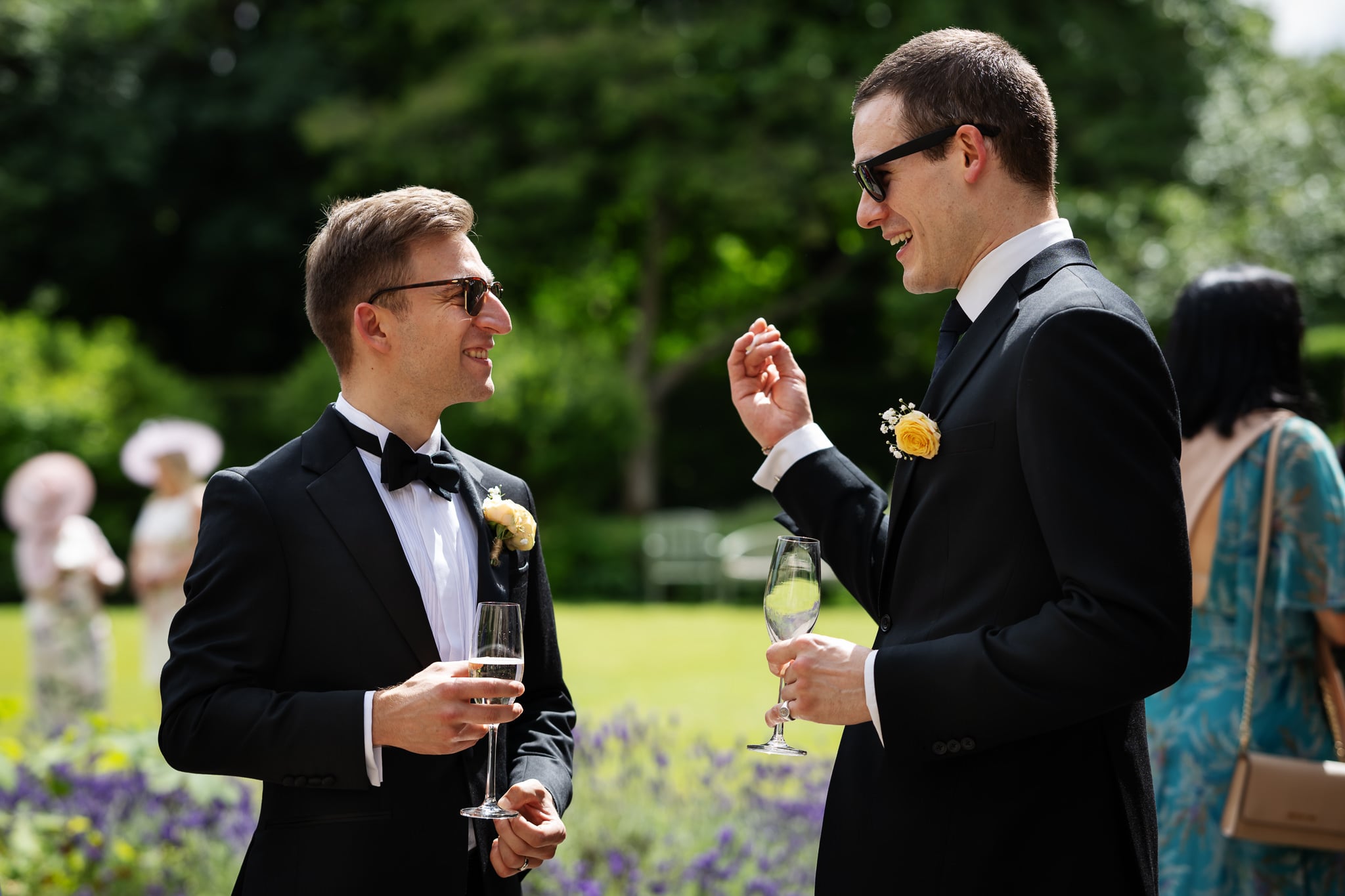 Two men in black suits and sunglasses, each holding a drink, smile and talk outdoors at a formal Notley Abbey event, with greenery, other guests, and the bride and groom in the background.