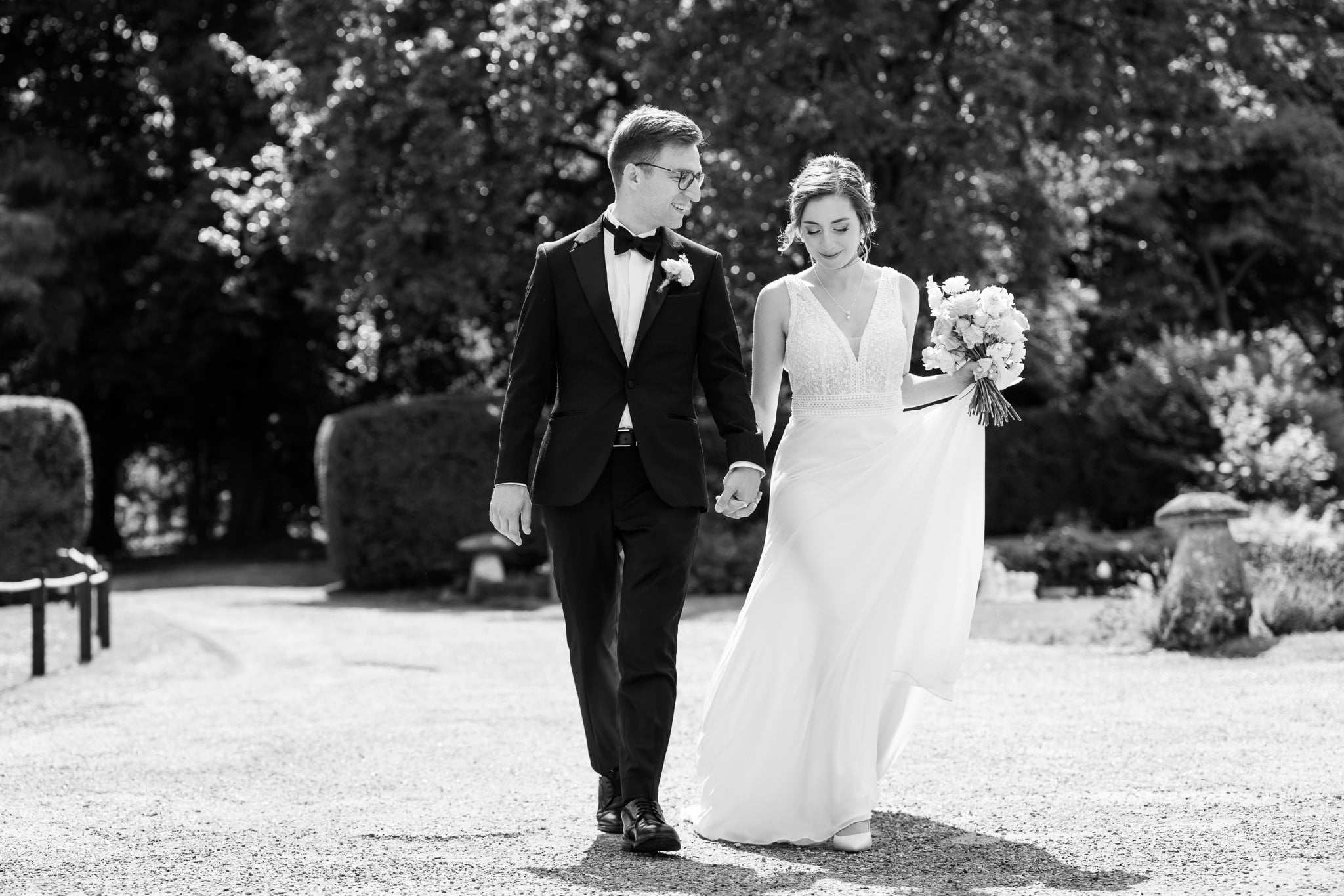 A bride and groom walk hand in hand outdoors at Notley Abbey, dressed in formal wedding attire, with trees and bushes in the Buckinghamshire countryside as their backdrop.