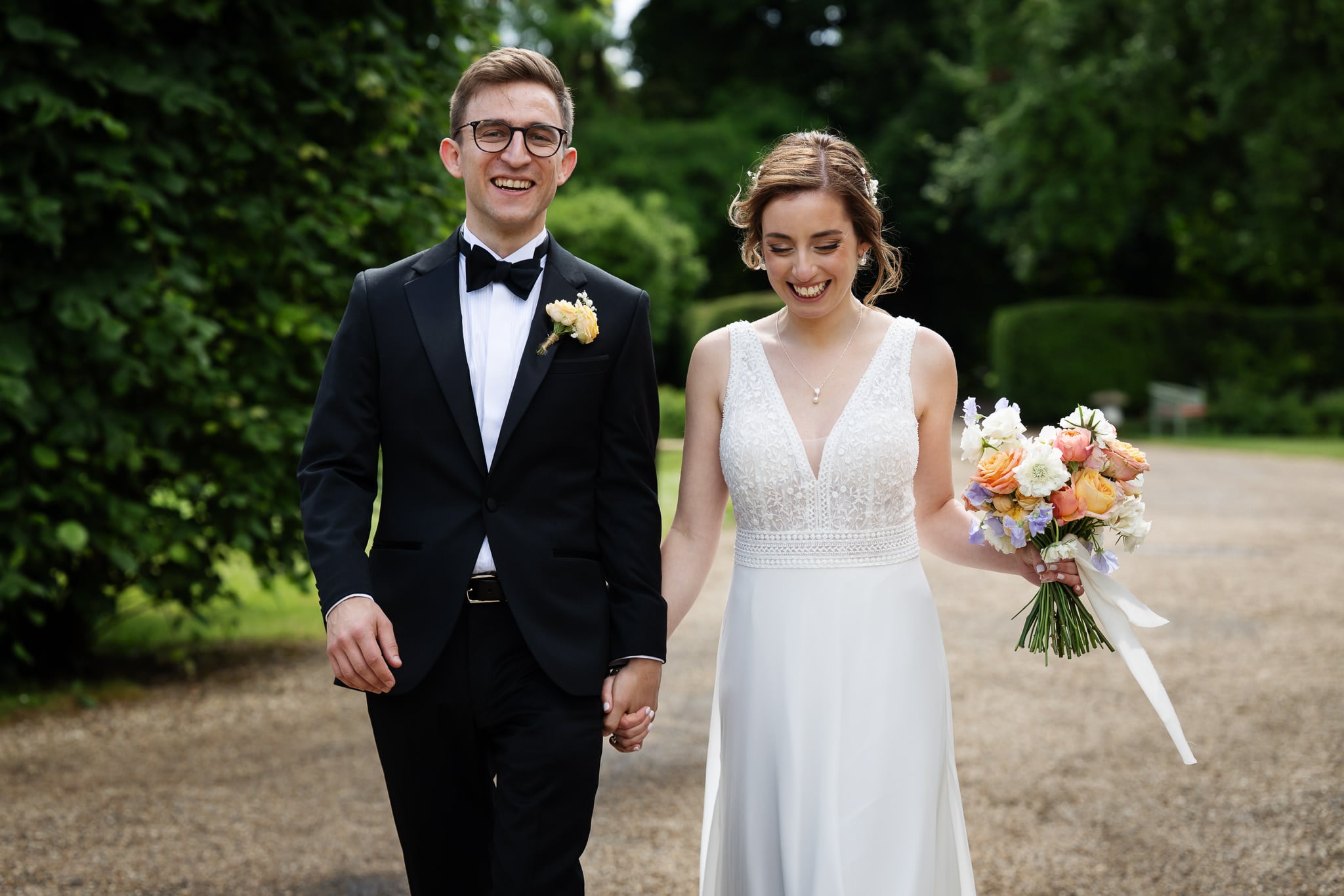 A smiling bride and groom walk hand in hand outdoors in Buckinghamshire. The groom wears a black tuxedo while the bride, holding a bouquet of flowers, looks radiant in her white dress.