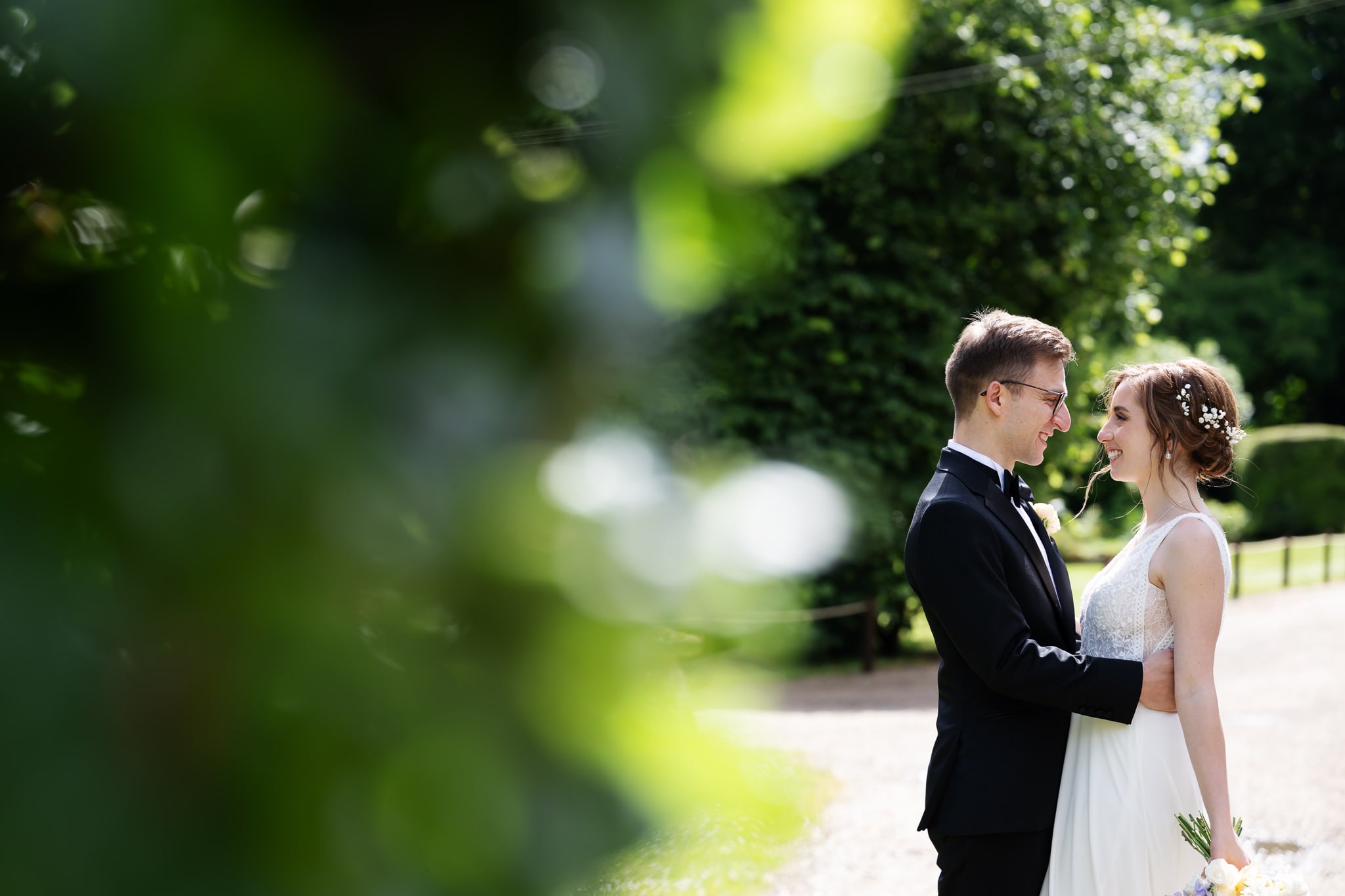 A bride and groom stand facing each other outdoors at their wedding, smiling, surrounded by greenery and sunlight.