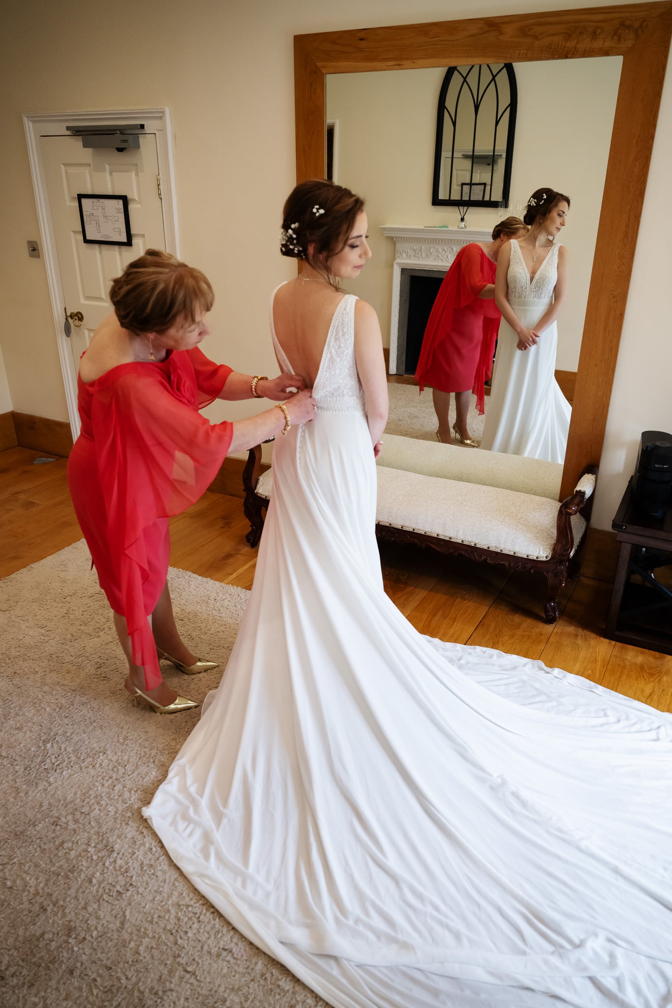A photographer captures a woman fastening the back of a bride's white wedding dress at Notley Abbey, as the bride stands before a large mirror with her train elegantly spread behind her.