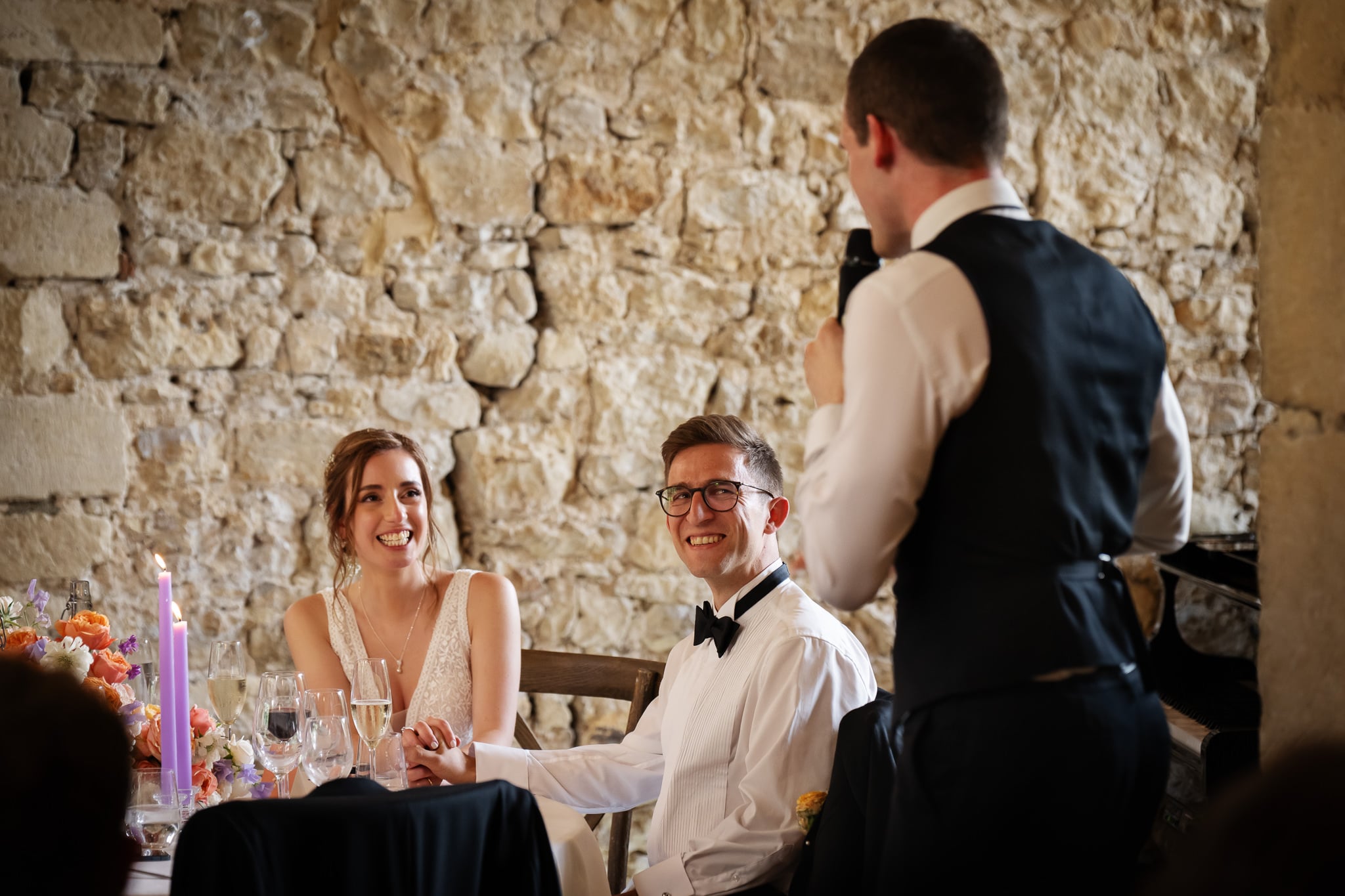 A newlywed couple sits at a table in Buckinghamshire, smiling and holding hands, while a man in a suit stands and speaks into a microphone in front of the happy groom and bride.