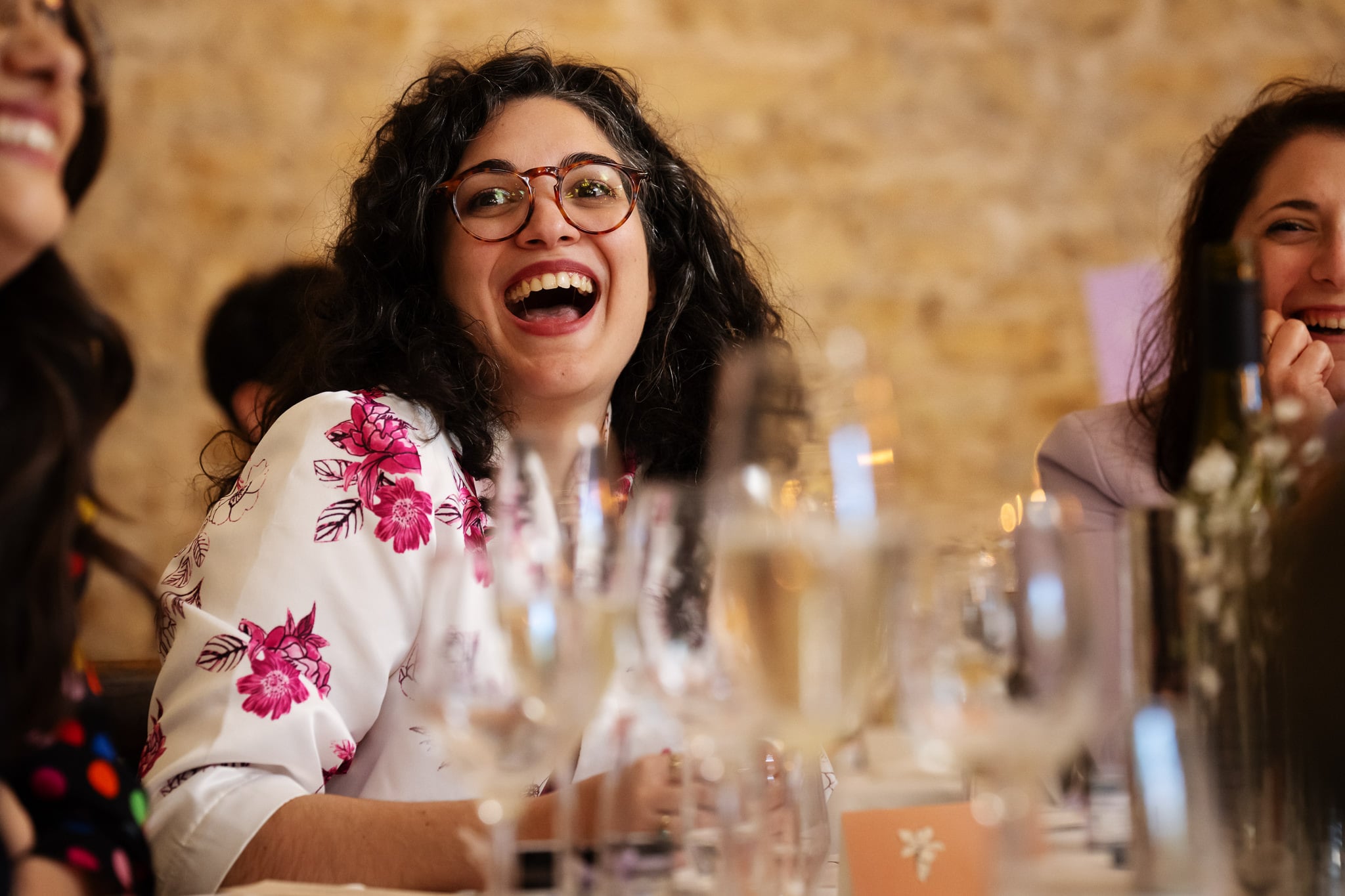 A woman with curly hair and glasses, possibly the bride, smiles and laughs while sitting at a table set with glasses and a wine bottle in Buckinghamshire.