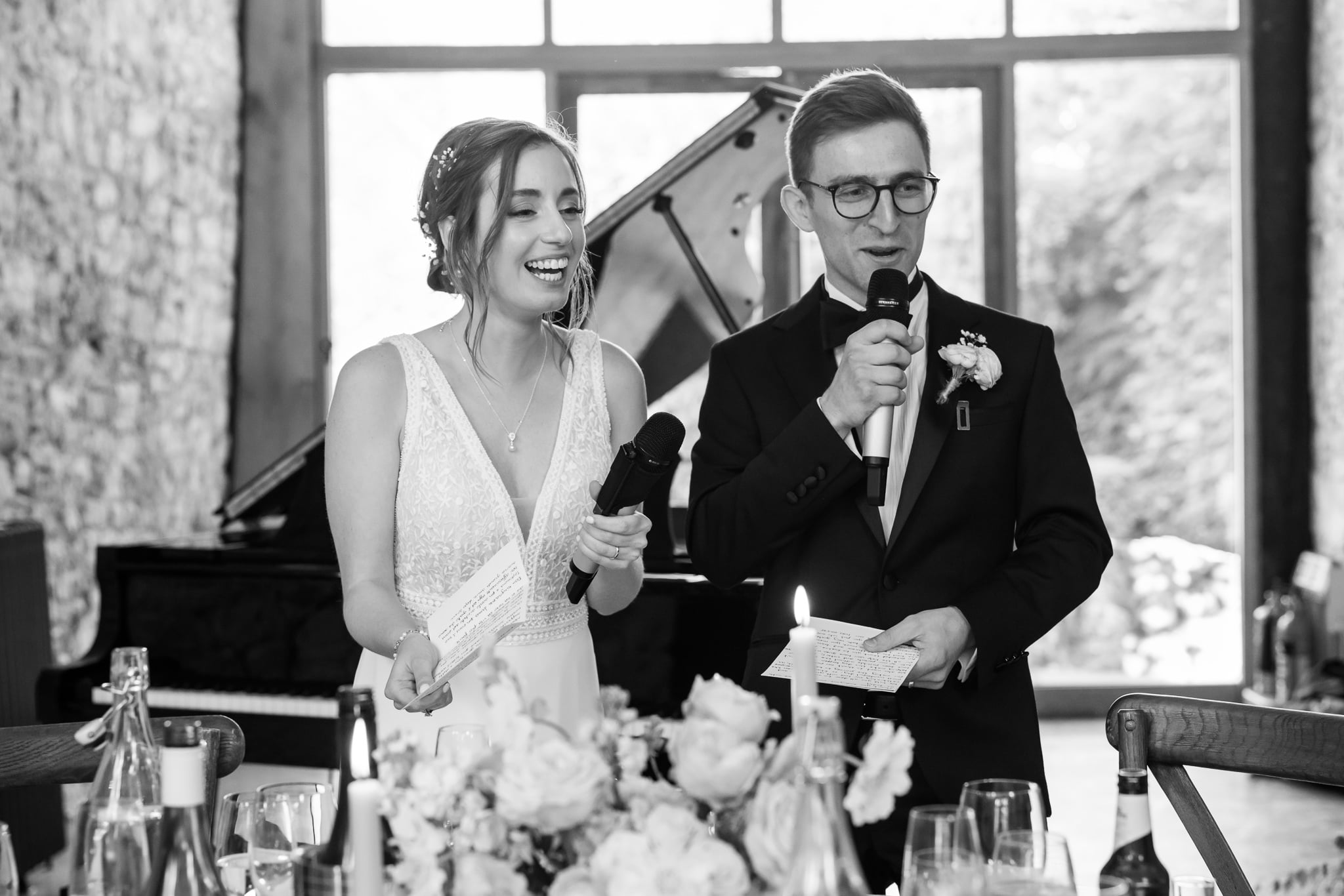 A bride and groom stand side by side holding microphones and notes, smiling as they give a speech at their wedding reception at Notley Abbey in Buckinghamshire. A piano is visible in the background.
