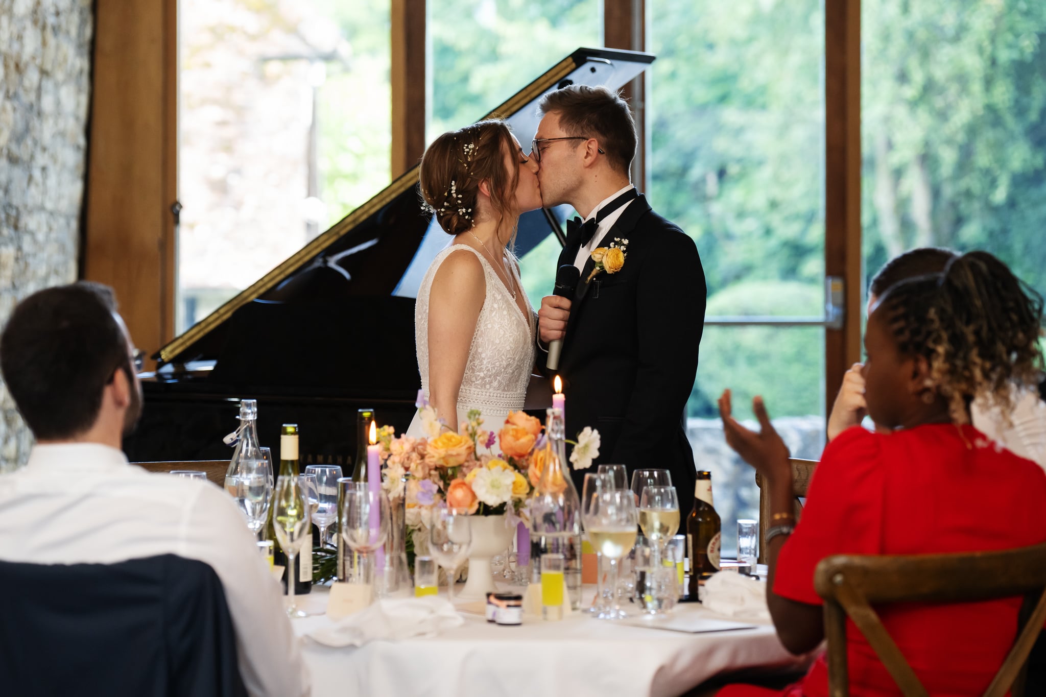 A bride and groom kiss beside a piano at a decorated table during their Notley Abbey wedding reception, as guests look on.
