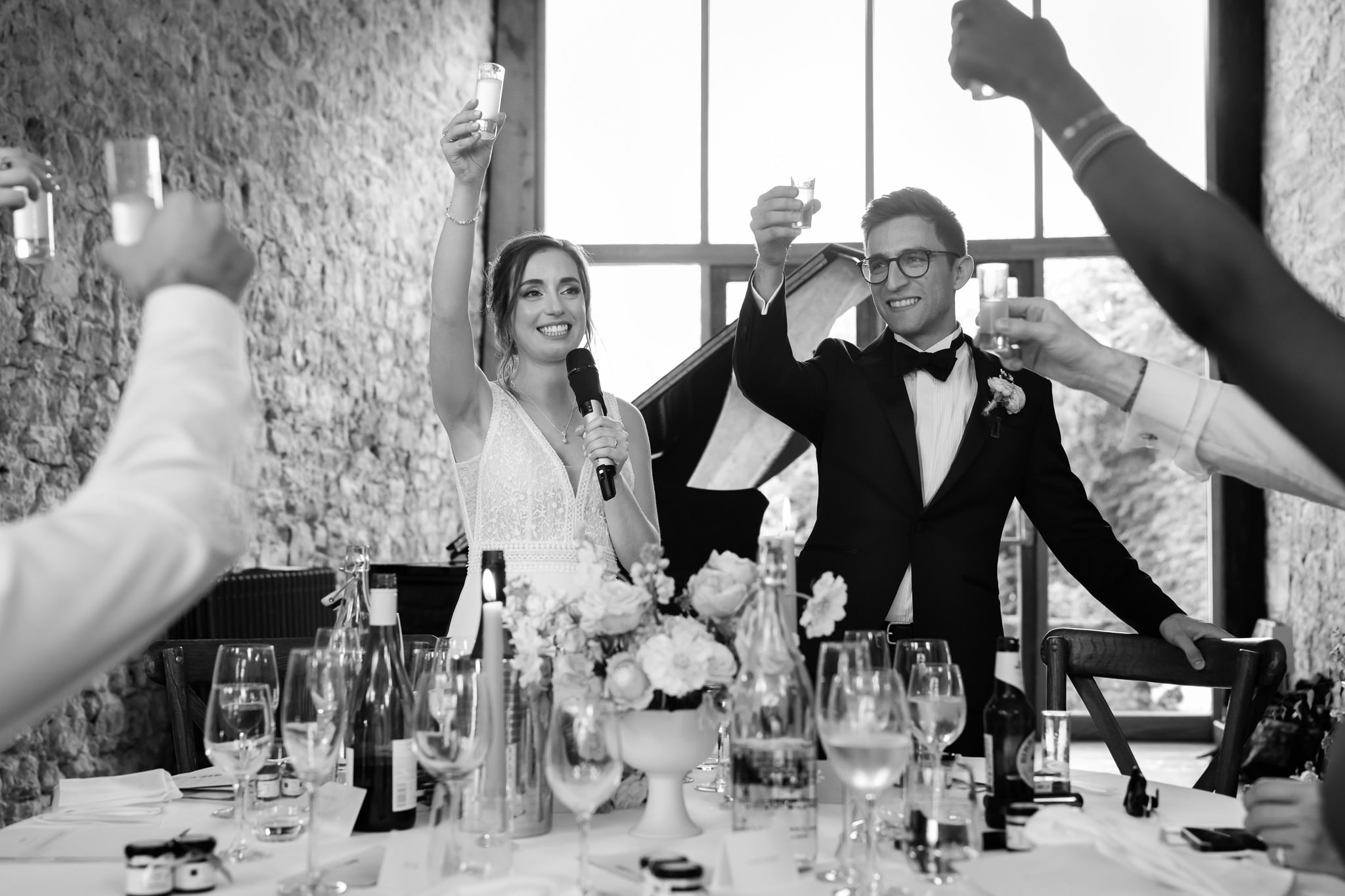 A bride and groom stand at a table raising a toast with guests in a bright, rustic Buckinghamshire venue, surrounded by flowers, glasses, and elegant table settings.
