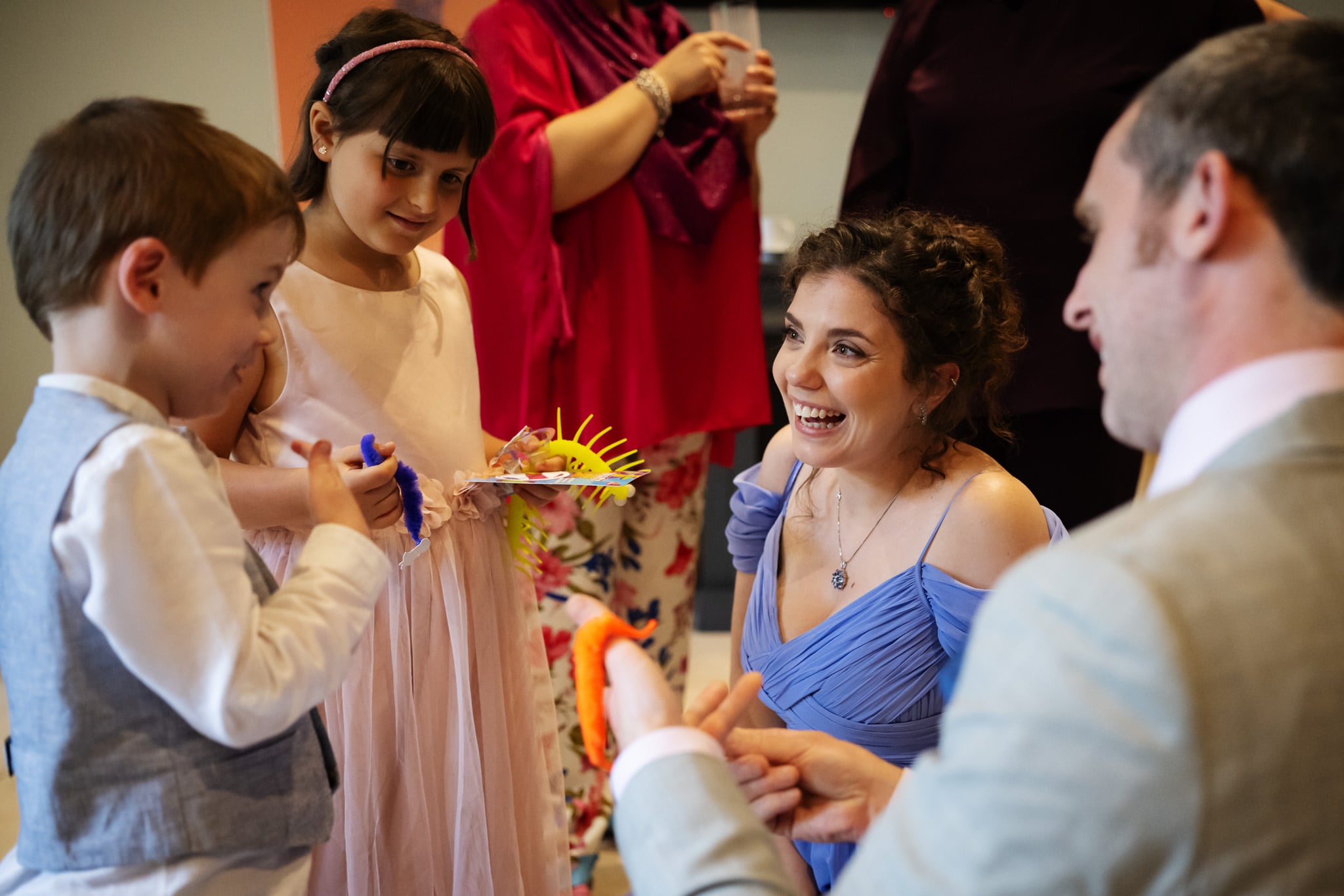 At a wedding at Notley Abbey, a woman in a lavender dress kneels and smiles at a young boy, while the groom, a girl in a pink dress, and other adults stand nearby at an indoor event.