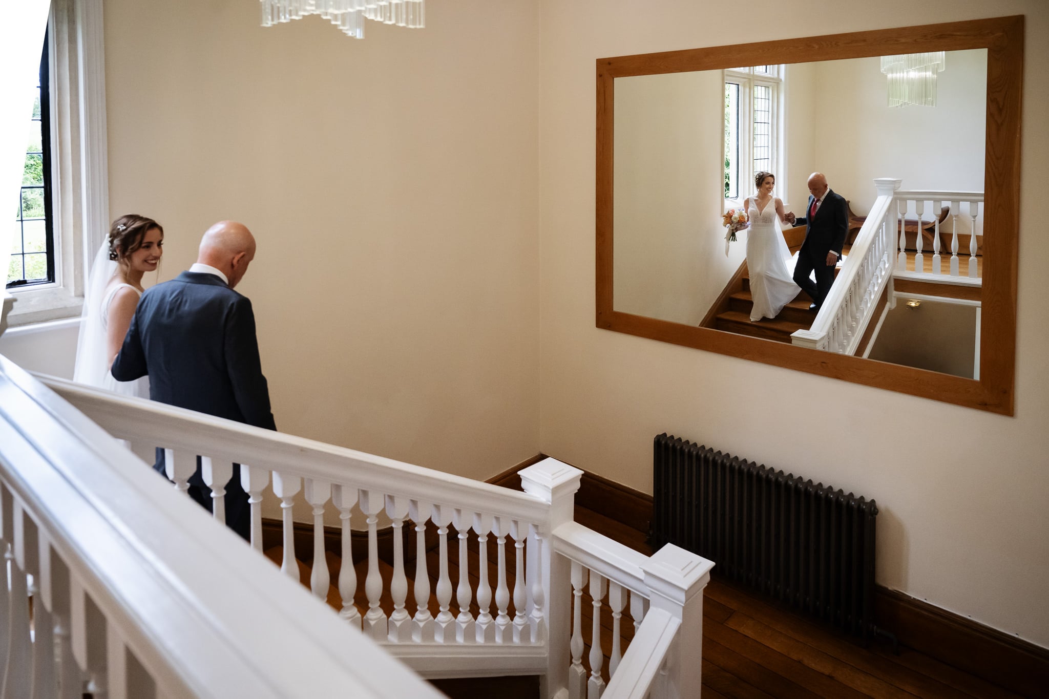 A bride and an older man, possibly her father, walk down a white staircase at a wedding, with their reflection visible in a large wall mirror.