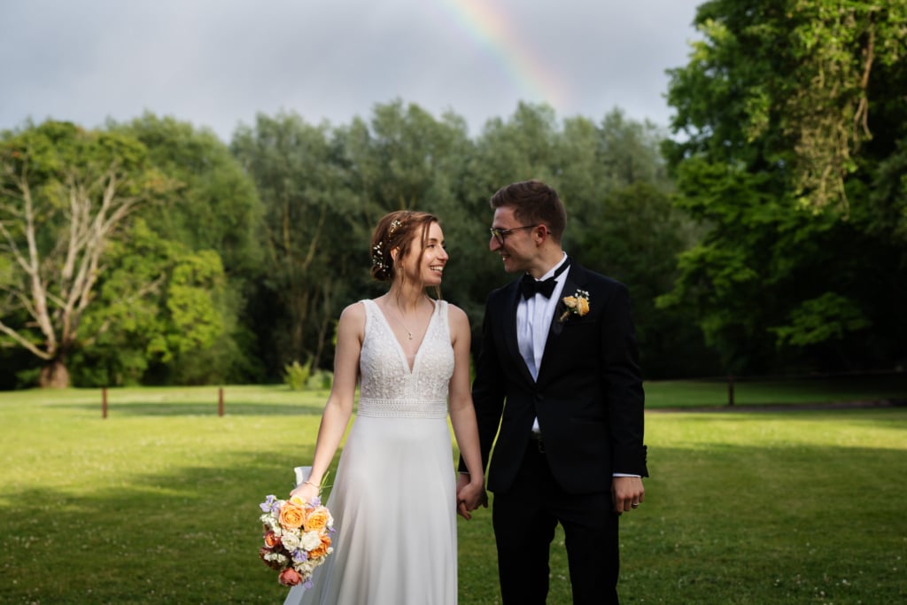 A bride and groom stand holding hands on the grass at Notley Abbey in Buckinghamshire, with trees and a faint rainbow behind them. The bride holds a bouquet, and both are smiling at each other.