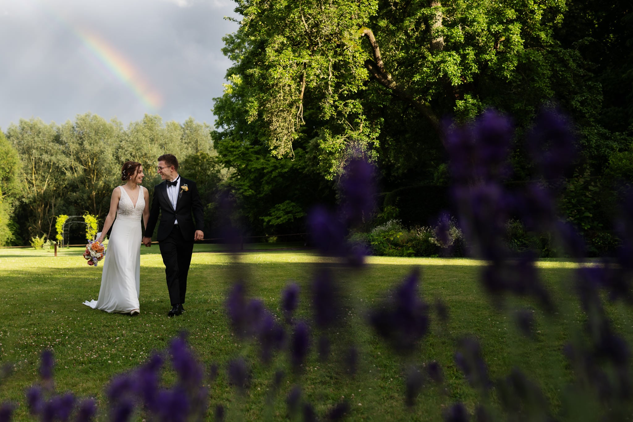 A bride and groom walk hand in hand on a grassy Buckinghamshire lawn, with purple flowers in the foreground and a faint rainbow arching across the sky behind them.