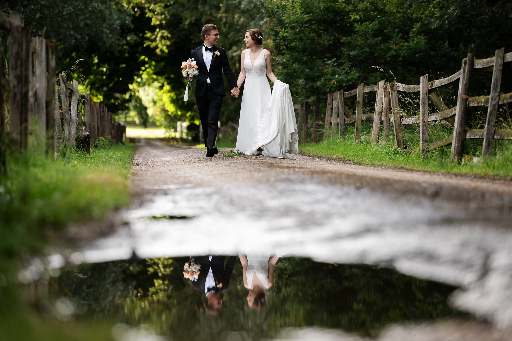 A bride and groom walk hand in hand down a country path at Notley Abbey, with a bouquet and their reflection visible in a puddle on the ground.