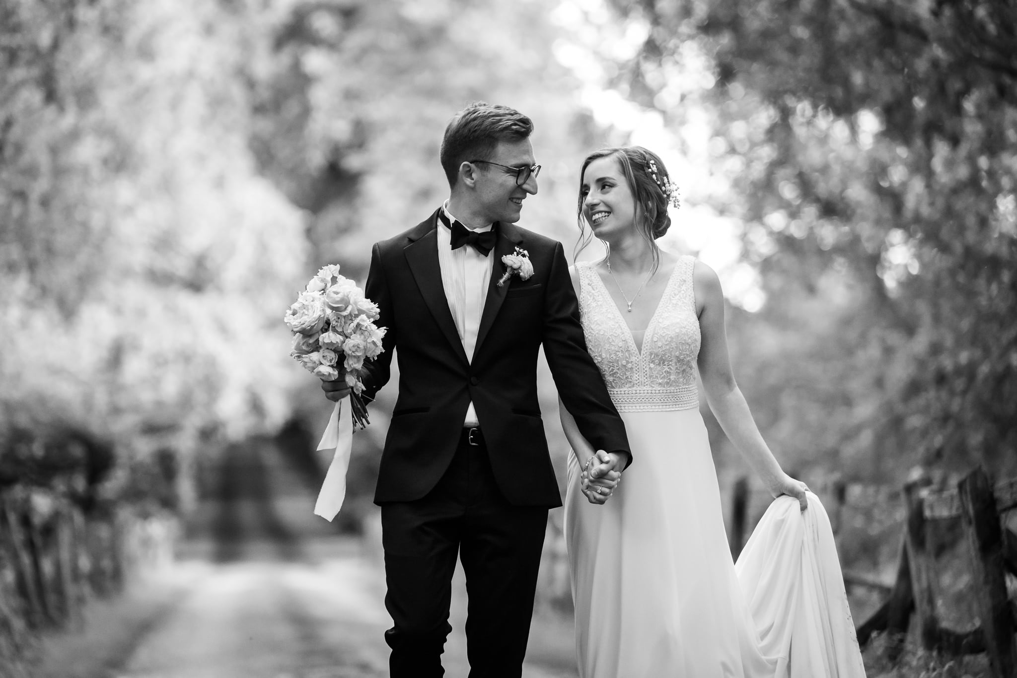 A bride and groom walk hand in hand down a tree-lined path at Notley Abbey in Buckinghamshire, with the groom holding a bouquet and both smiling at each other.