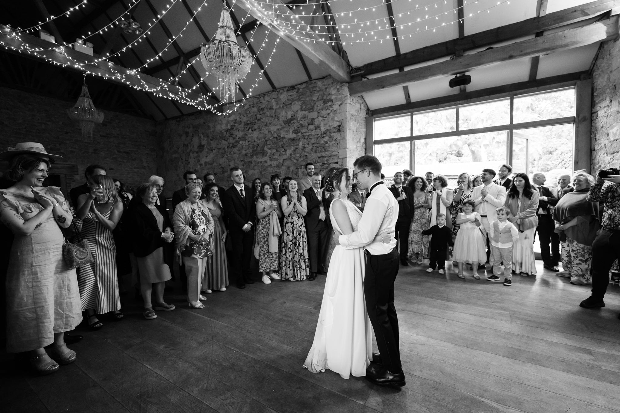 A bride and groom share their first dance at Notley Abbey, a rustic Buckinghamshire venue, surrounded by smiling guests as string lights hang from the ceiling.