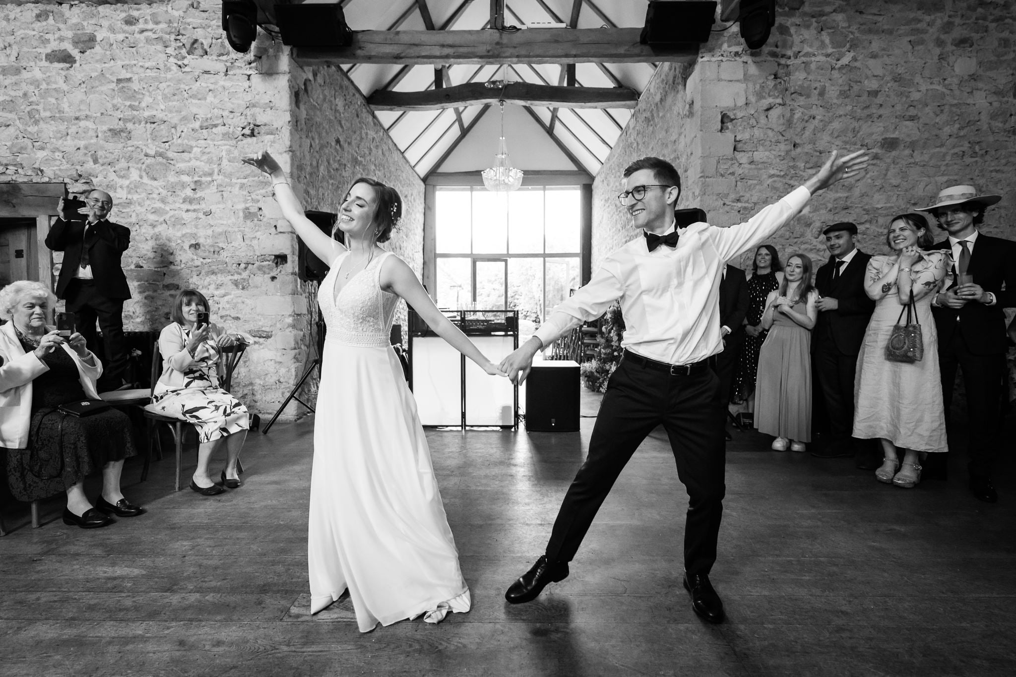 A bride and groom hold hands and dance in the rustic hall at Notley Abbey as guests watch and take photos.
