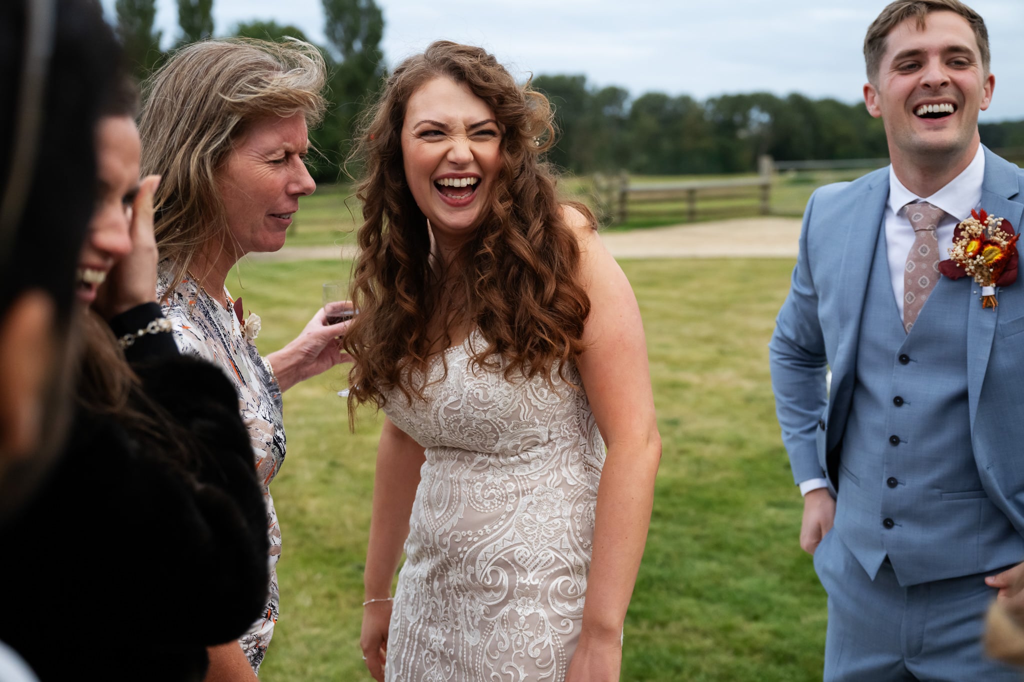 A woman in a white patterned dress laughs with others outdoors at an Aylesbury wedding venue, while a man in a light blue suit stands nearby, also smiling.