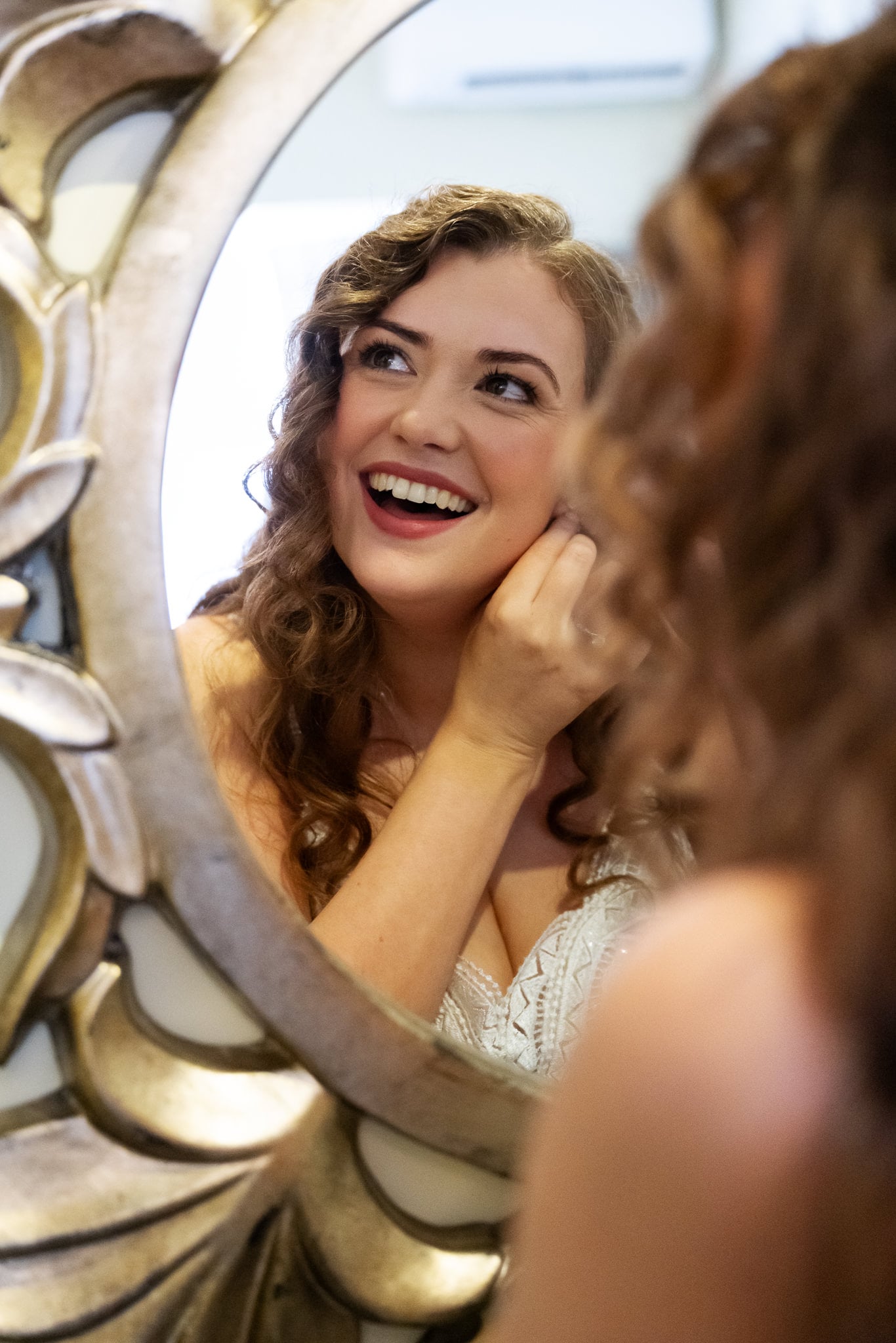 A woman in a white dress smiles while putting on an earring at an Aylesbury wedding venue, her reflection visible in an ornate mirror.