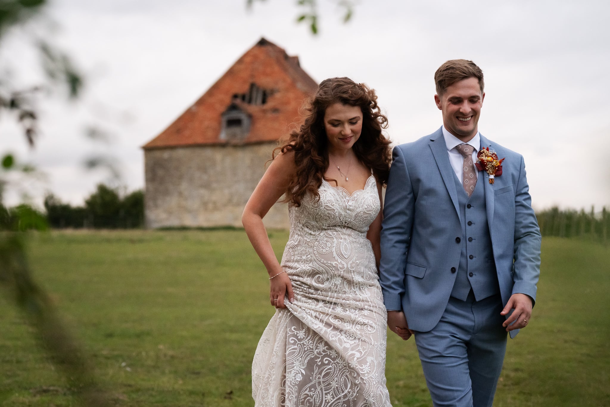 A bride in a lace dress and a groom in a light blue suit walk hand in hand outdoors at Notley Tythe Barn, with an old stone building highlighting this charming barn wedding venue.