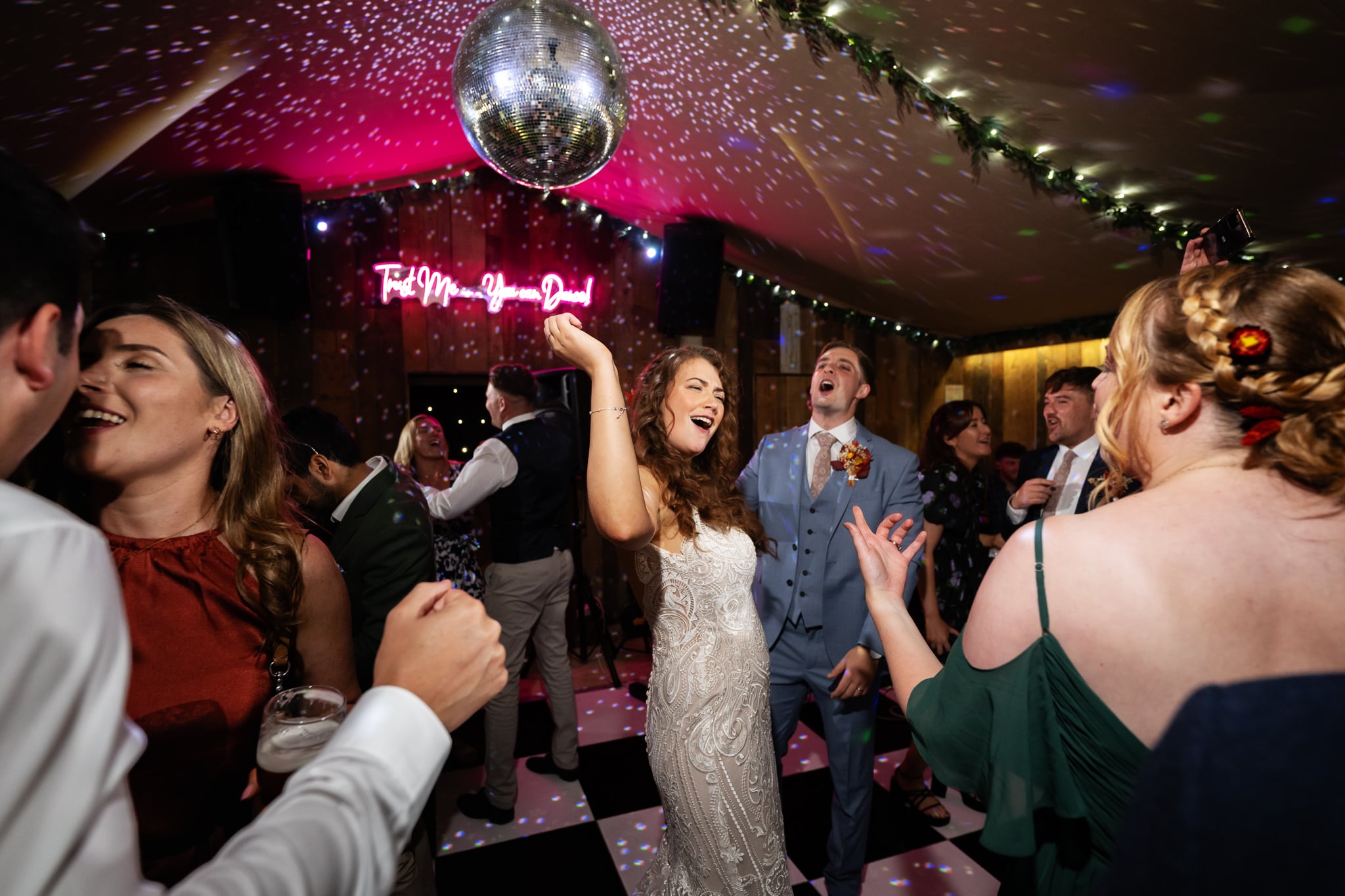 A bride in a white dress dances joyfully under a disco ball at Notley Tythe Barn, surrounded by guests on a checkered dance floor at a lively indoor wedding celebration.