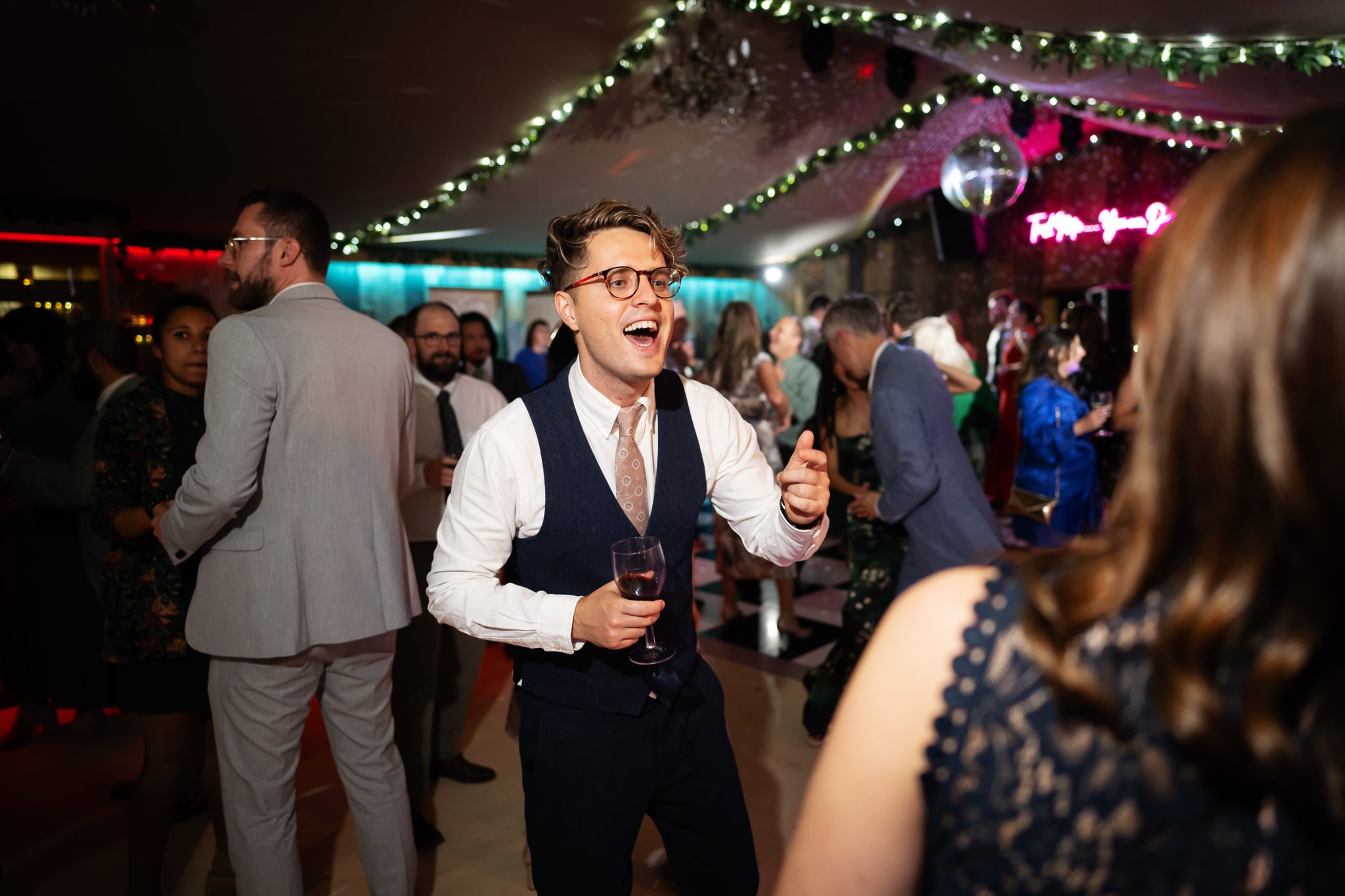 A person in a navy vest and glasses laughs while holding a drink on a crowded, decorated dance floor at a barn wedding venue.