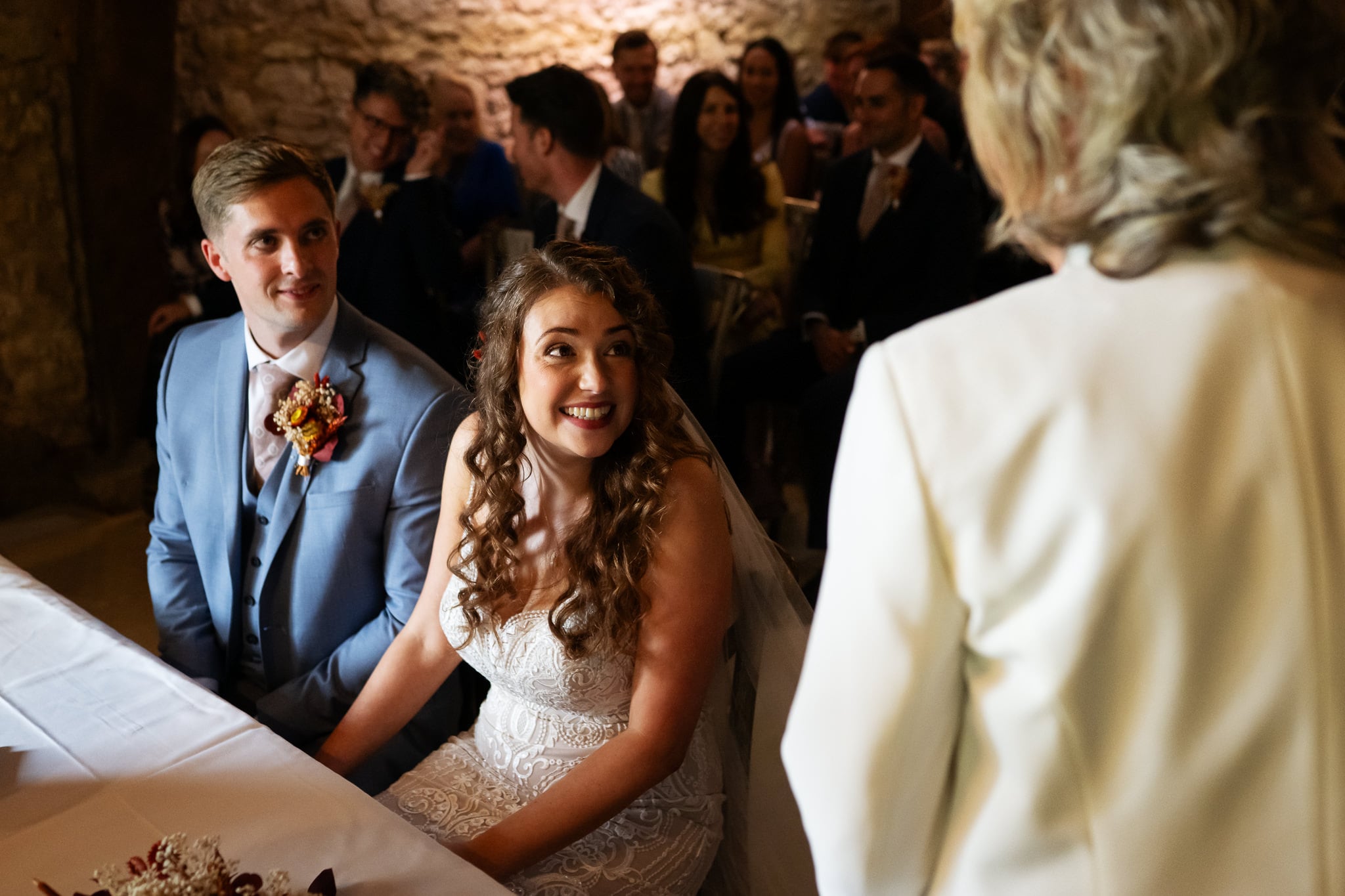 A bride and groom sit at a table during their wedding ceremony at Notley Tythe Barn, a stunning barn wedding venue, facing the officiant in white while guests are seated in the background.