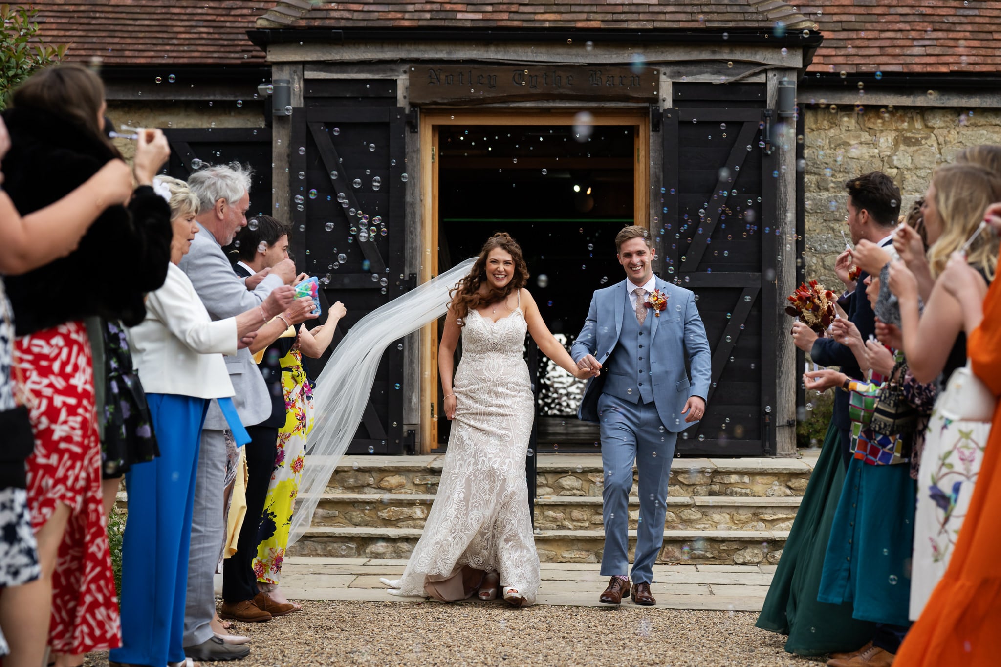 A bride and groom walk out of Notley Tythe Barn, smiling and holding hands, as guests blow bubbles and celebrate on either side—a perfect moment captured by the photographer at this beautiful barn wedding venue.