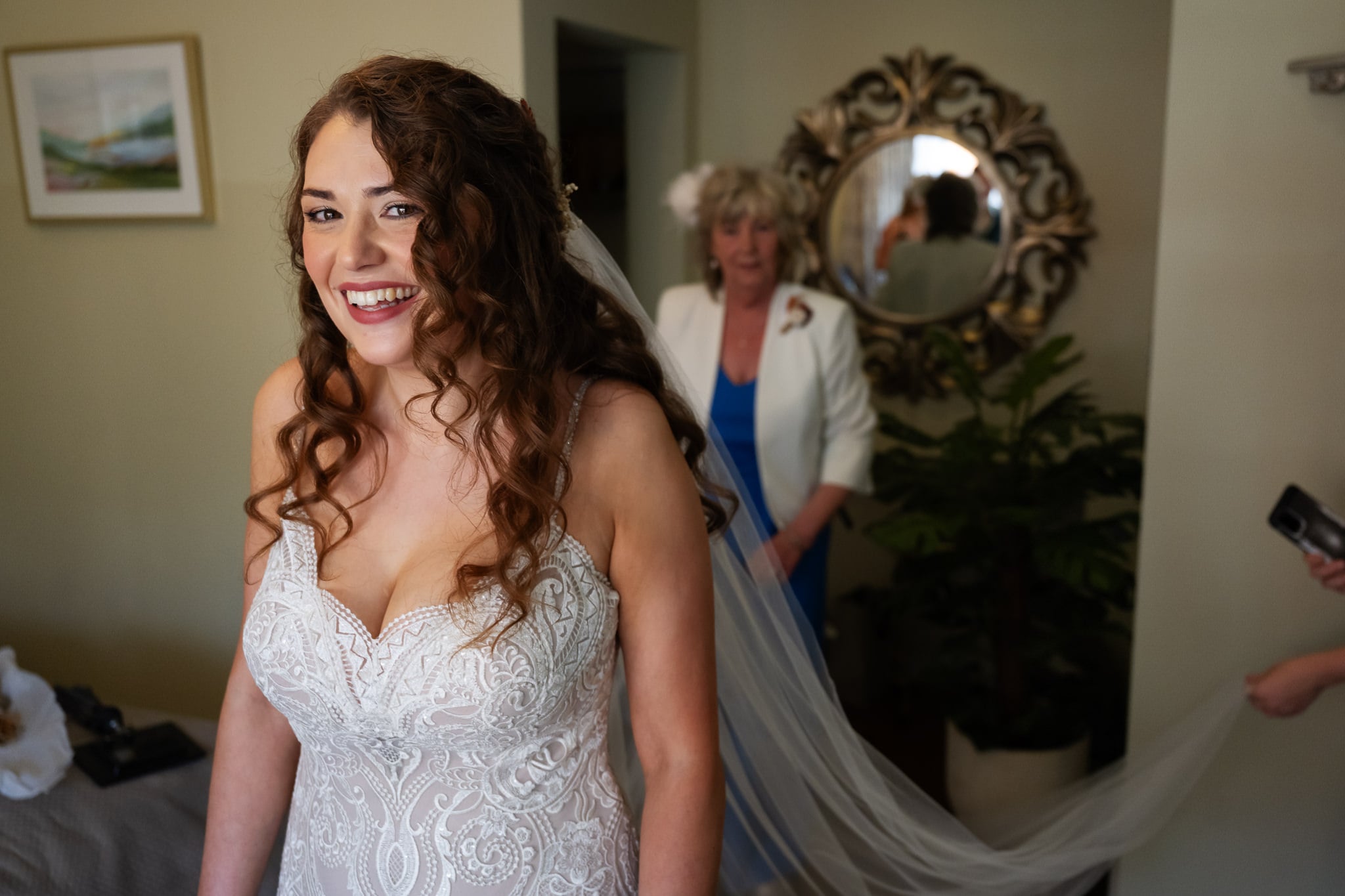 A smiling bride in a lace wedding dress stands in a room at a charming barn wedding venue, while another woman in the background adjusts her veil as the photographer captures the moment.