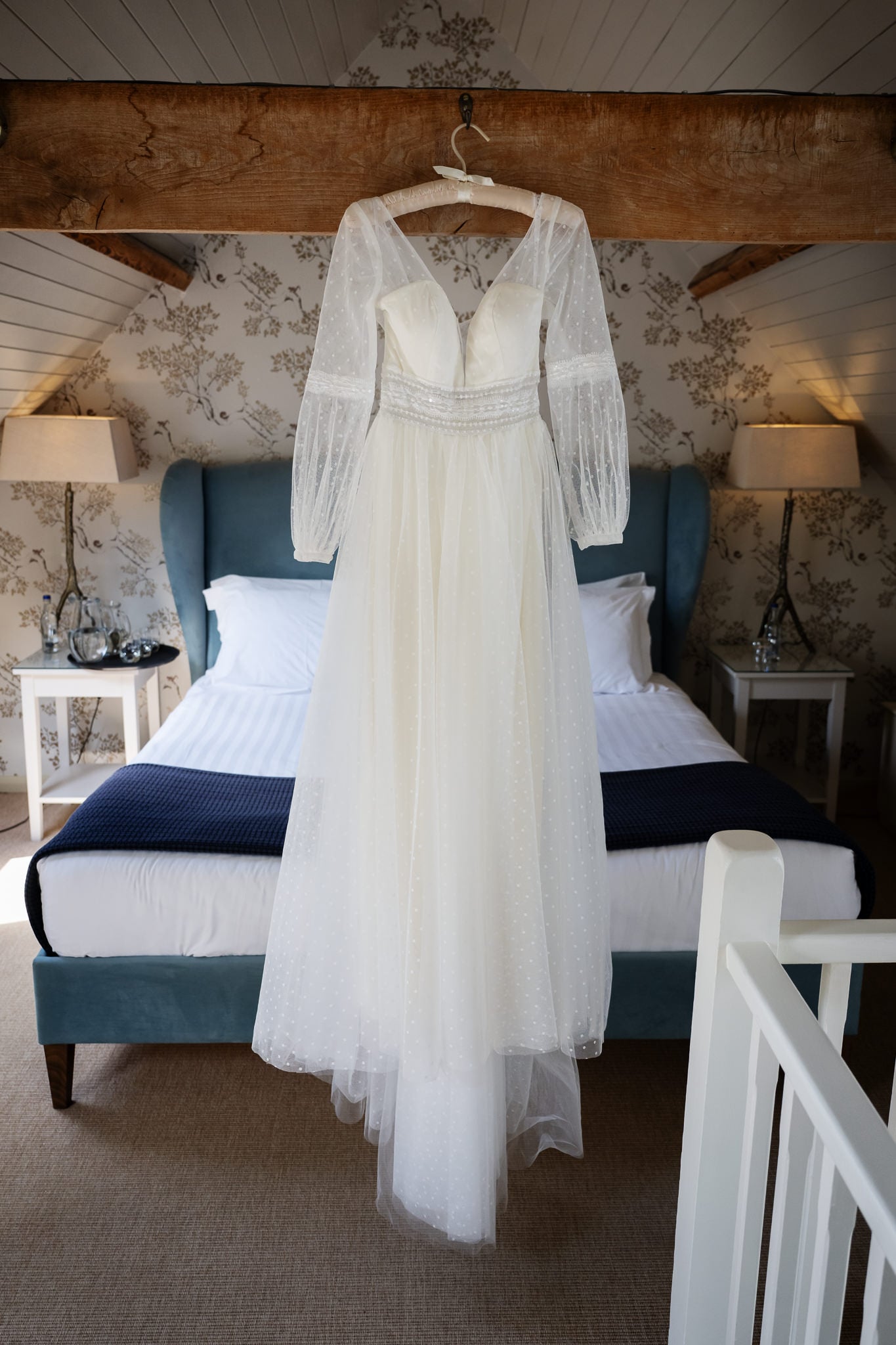 A white, long-sleeved wedding dress hangs on a wooden beam above a neatly made bed in a decorated bedroom, perfect for a Bride getting ready at a charming Cambridgeshire wedding venue.