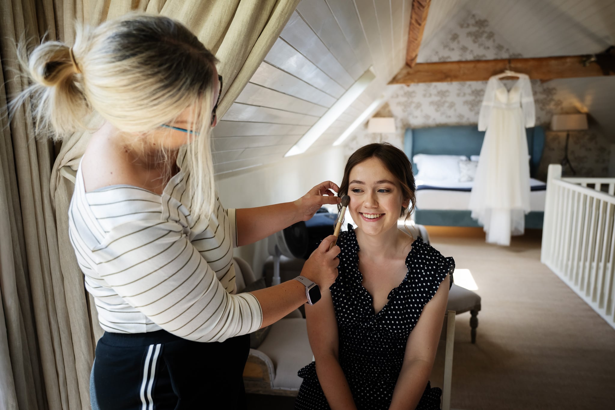 A Bride has her hair styled by another woman in a bright attic room. A white dress hangs in the background on the wall near a bed, evoking the charm of a Cambridgeshire wedding venue.