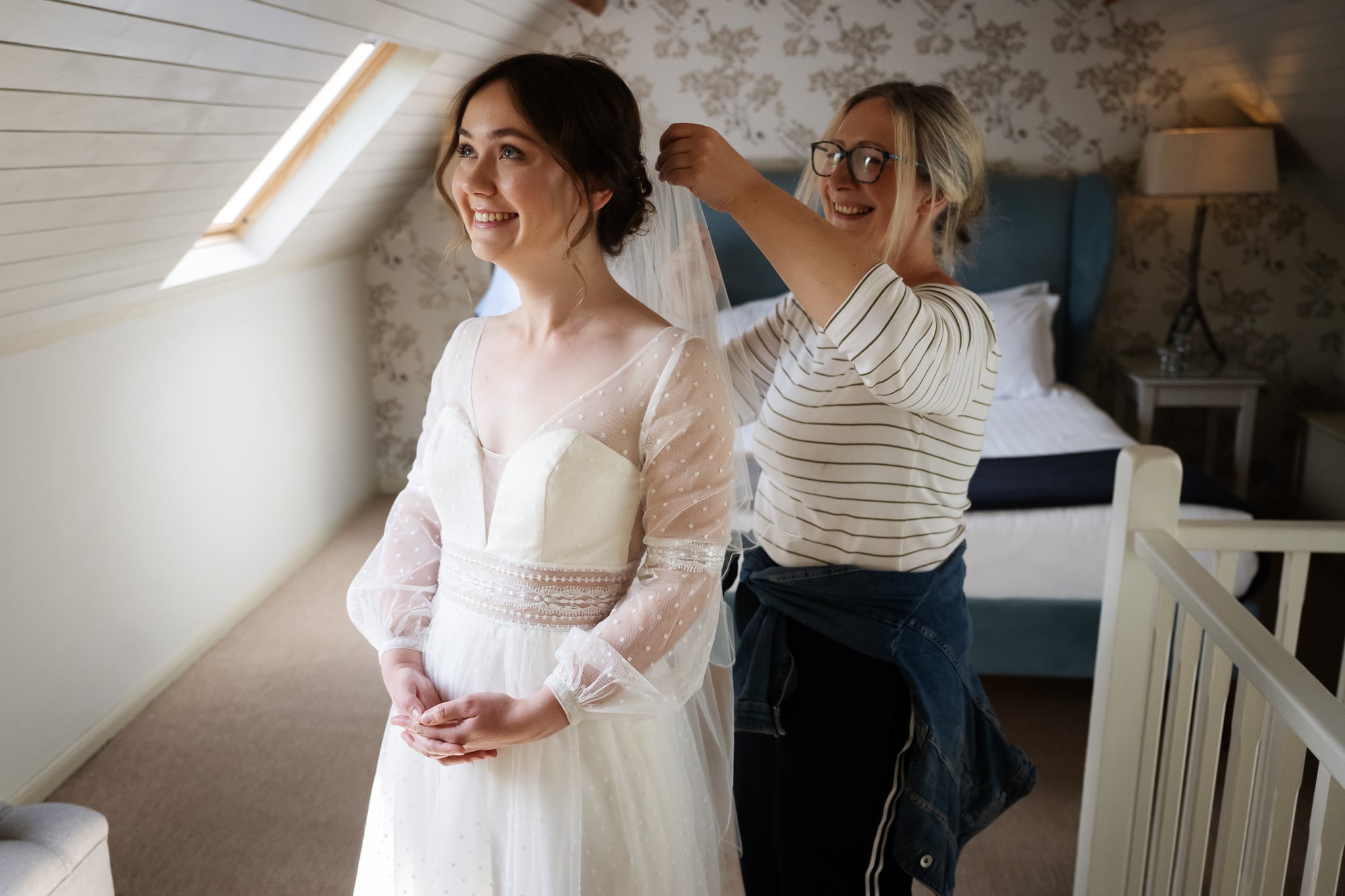 A woman in a white wedding dress smiles as another woman adjusts her veil in a softly lit bedroom with floral wallpaper at a charming Cambridgeshire wedding venue.