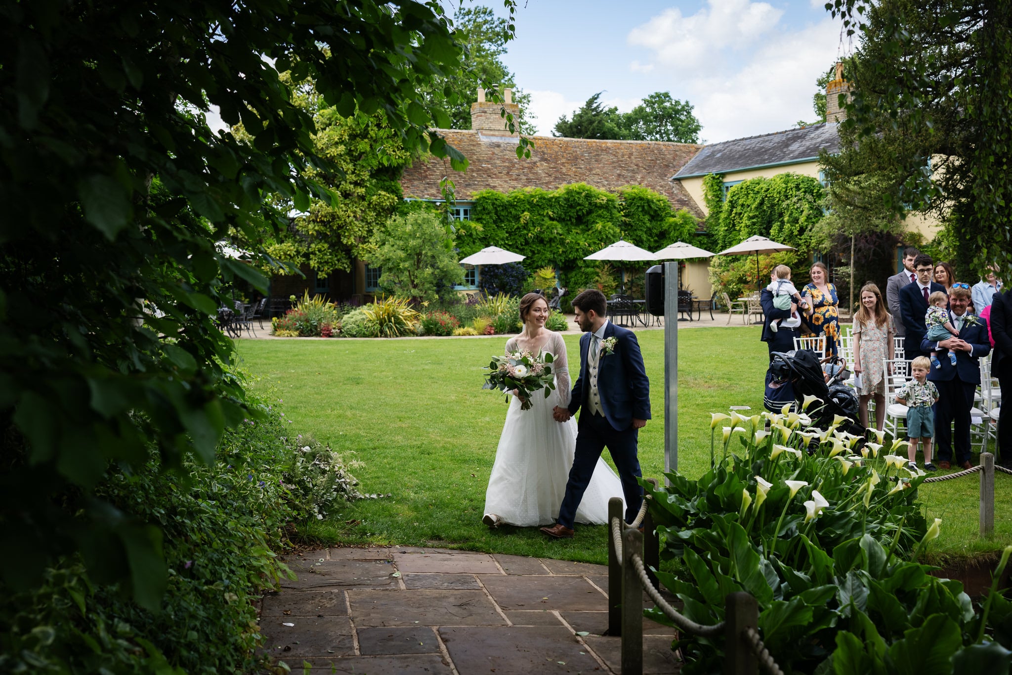 A bride and groom walk hand in hand across a garden lawn at a Cambridgeshire wedding venue as guests watch and take photos; outdoor seating and umbrellas are visible in the background.