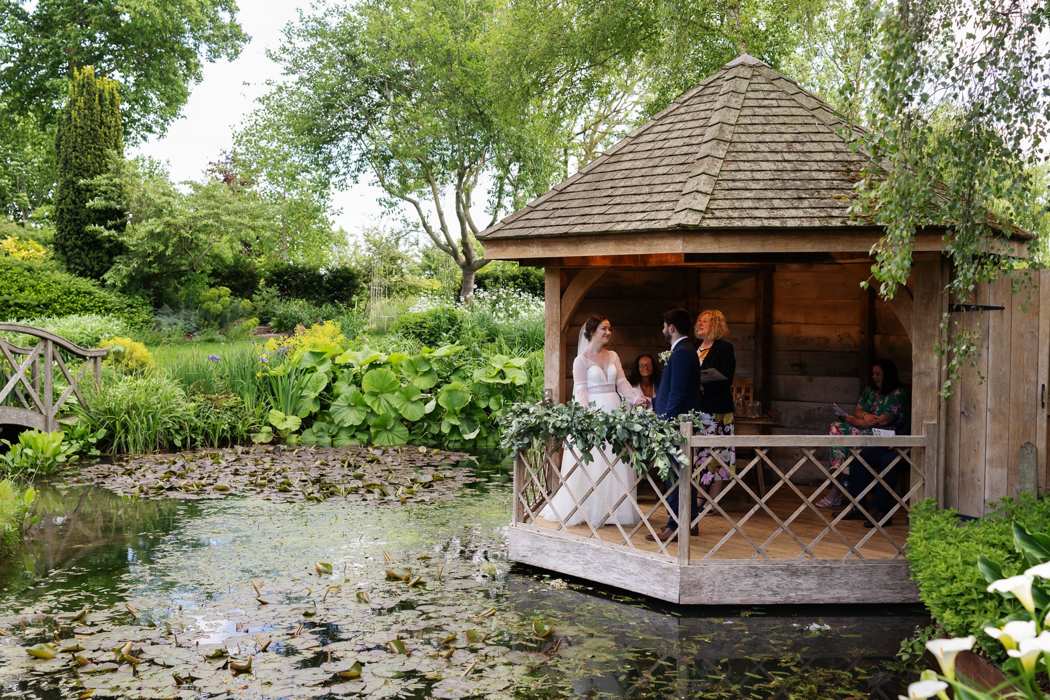 A bride and groom stand together inside a wooden gazebo by a pond at this charming Cambridgeshire wedding venue, surrounded by greenery, with a few seated guests nearby.