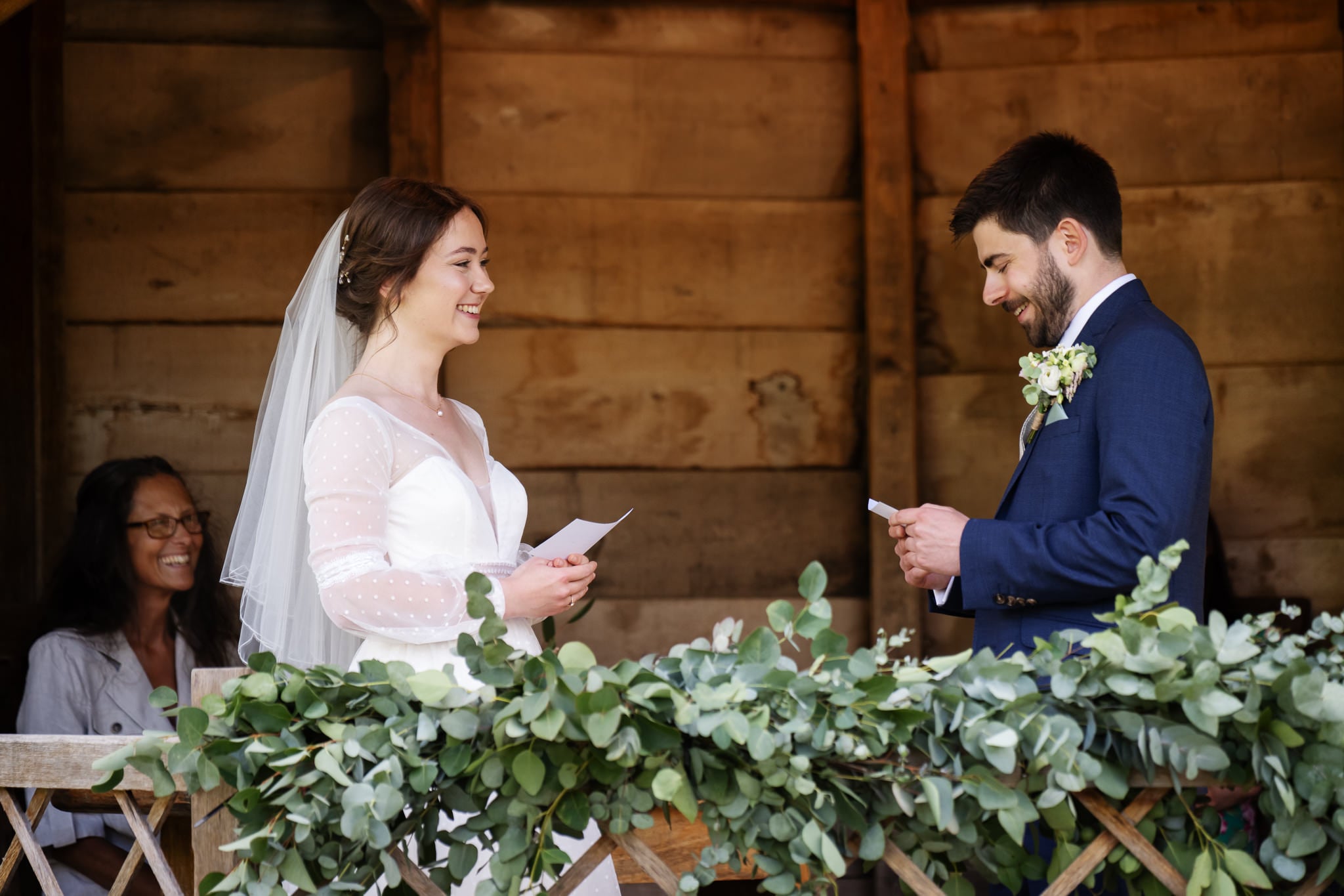 A bride and groom exchange vows while smiling at each other, standing in front of greenery with a wooden backdrop at a beautiful Cambridgeshire wedding venue. A woman sits behind them, watching and smiling.